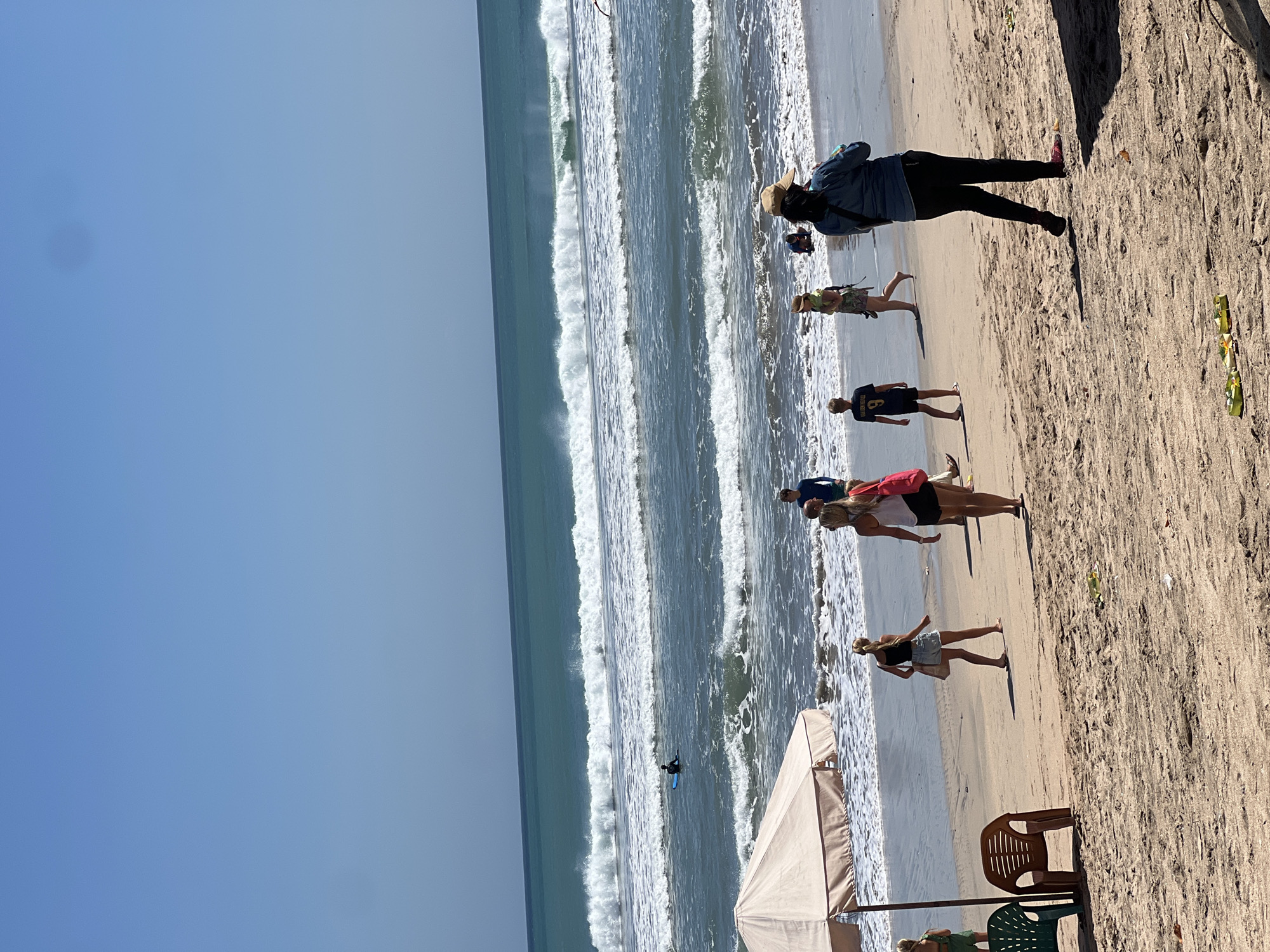 A wide shot captures a sunny day at Kuta Beach, Indonesia, with a clear blue sky overhead. The ocean features several layers of breaking waves, with a lone surfer visible on a surfboard in the distance. Along the sandy shoreline, a group of people are enjoying the beach. To the left, a large, beige beach umbrella shades two empty plastic chairs, one green and one brown. Several individuals are walking on the beach, including a woman in a black tank top and denim shorts, another woman in a white tank top and black shorts carrying a pink bag, a woman in a hat wearing a green dress, a young boy in a black t-shirt with the number "9" on it, and a woman wearing a denim jacket and hat facing away from the camera. A person in a blue shirt is visible further down the beach, partially obscured by others. The sand is marked with footprints and some scattered debris.