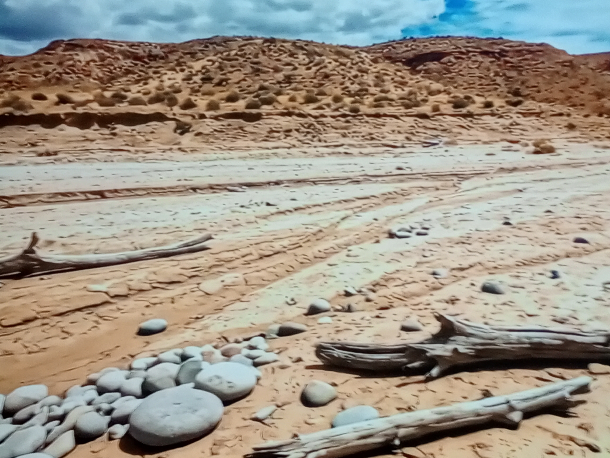The image depicts a dry, sandy riverbed with a backdrop of rugged, eroded hills under a cloudy sky. Several pieces of driftwood are scattered across the riverbed, along with a collection of smooth, rounded stones. Tire tracks are visible in the sand, indicating recent vehicular activity. The overall scene is one of a desolate, arid landscape.