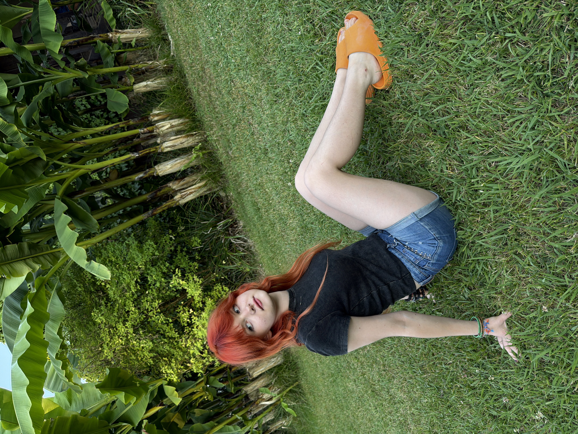 A young woman with long, bright red hair lies on her back in a grassy area, with a backdrop of lush green foliage and what appear to be banana trees. She is wearing a dark t-shirt and denim shorts, with orange slides on her feet. Her body is positioned in a relaxed pose, with her legs bent and one arm extended upwards, her fingers reaching out. She is looking towards the camera with a soft expression. The lighting suggests it is daytime, with the sun illuminating the scene. The presence of banana trees and the overall verdant environment point to a warm climate.