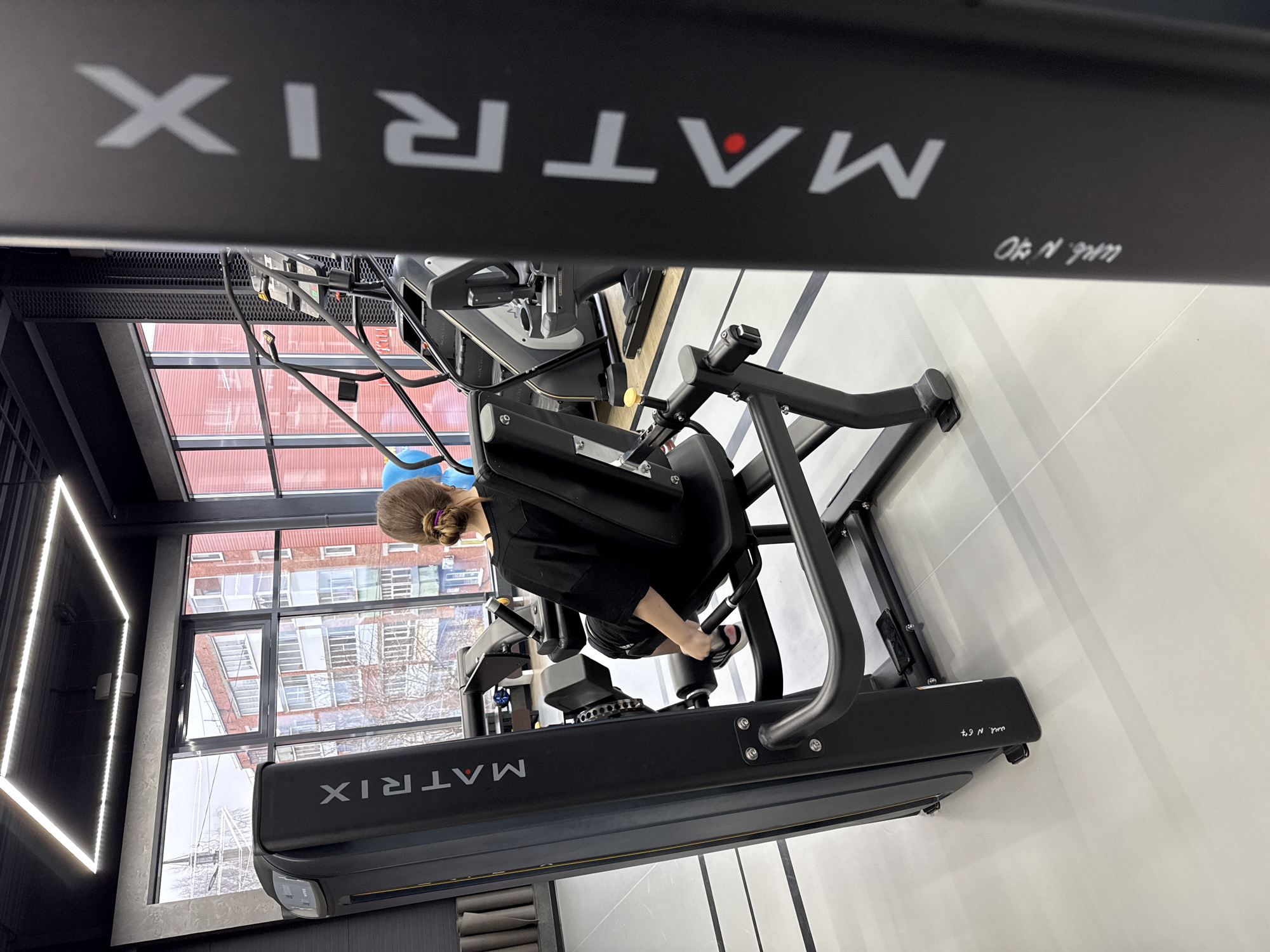 A woman wearing a black shirt and shorts is using a Matrix fitness machine in a gym with large windows, appearing to be in Votkinsk, Russia. The gym is well-lit, and outside the windows are buildings.