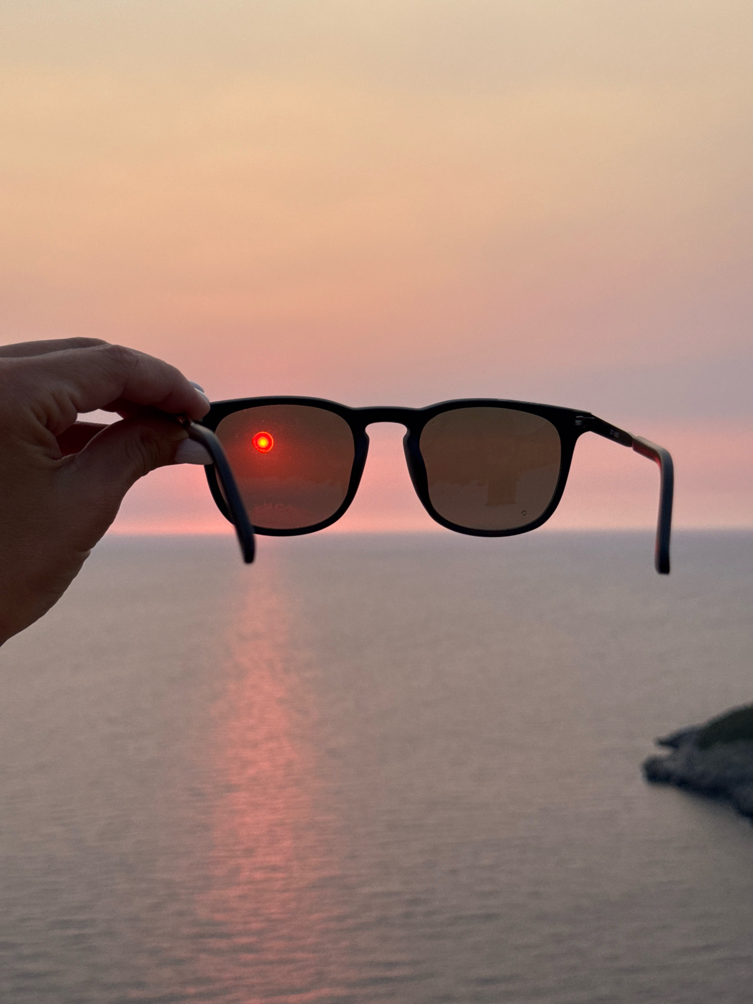 A hand holds a pair of black-rimmed sunglasses in front of a sunset over the Adriatic Sea, near Zagora, Montenegro. The setting sun, a vibrant red orb, is reflected in the left lens of the sunglasses, creating a fiery halo. The sky transitions from a soft pink to a pale orange, and the sea below is calm with a shimmering reflection of the sunset. The dark silhouette of a rocky coastline is visible in the lower right corner of the image.