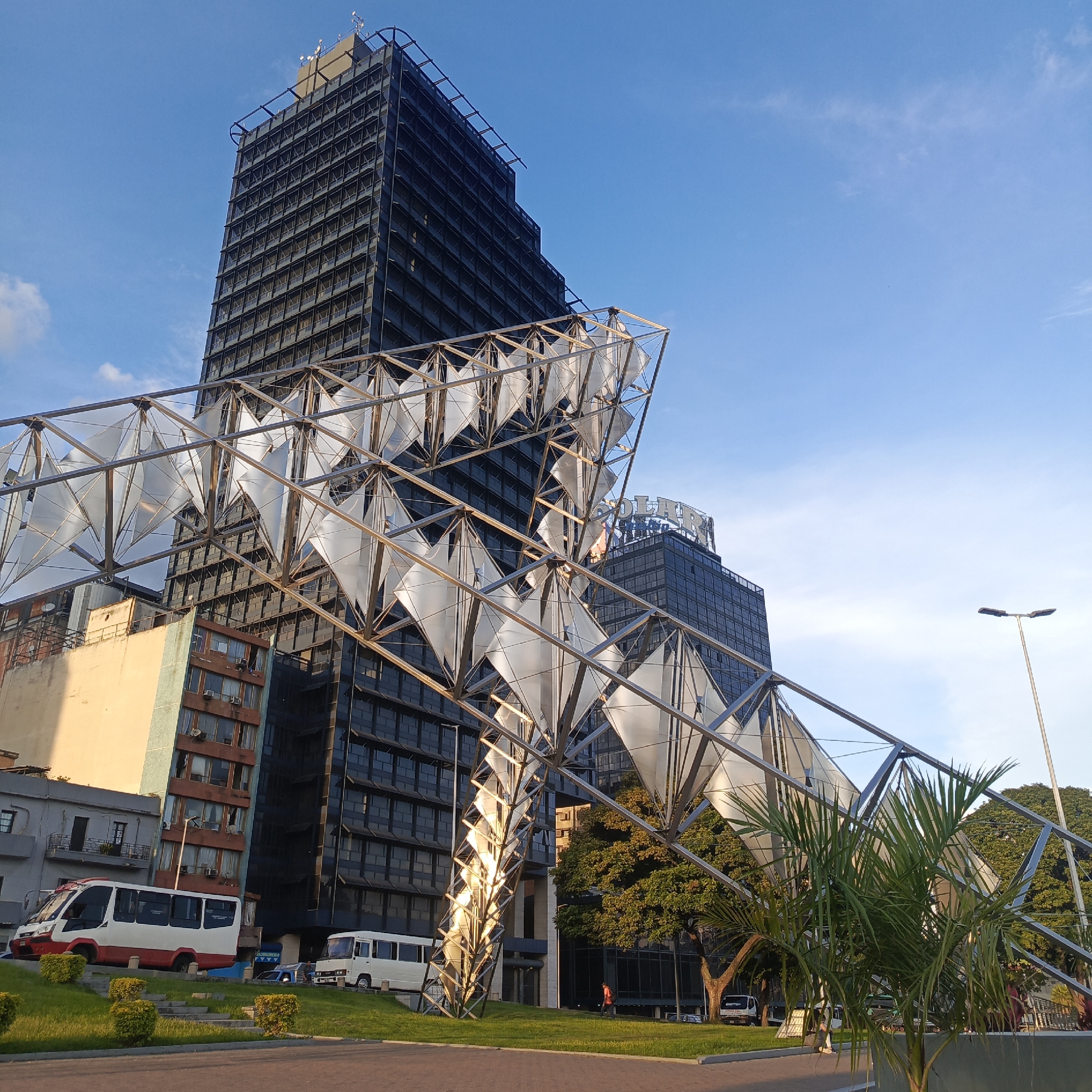 The image captures a public space in Caracas, Venezuela, featuring a modern, angular metallic sculpture with translucent diamond-shaped panels in the foreground. Behind the sculpture, a tall, dark glass-facade skyscraper dominates the background, with a lighter-colored building to its left. The sky is a clear, bright blue with a few wispy clouds. In the lower left corner, a white and red bus is parked on a grassy area with neatly trimmed bushes. Another white bus is visible further back, near a street. A street lamp is present on the right side of the frame. The overall scene suggests an urban environment with contemporary architecture and public art.