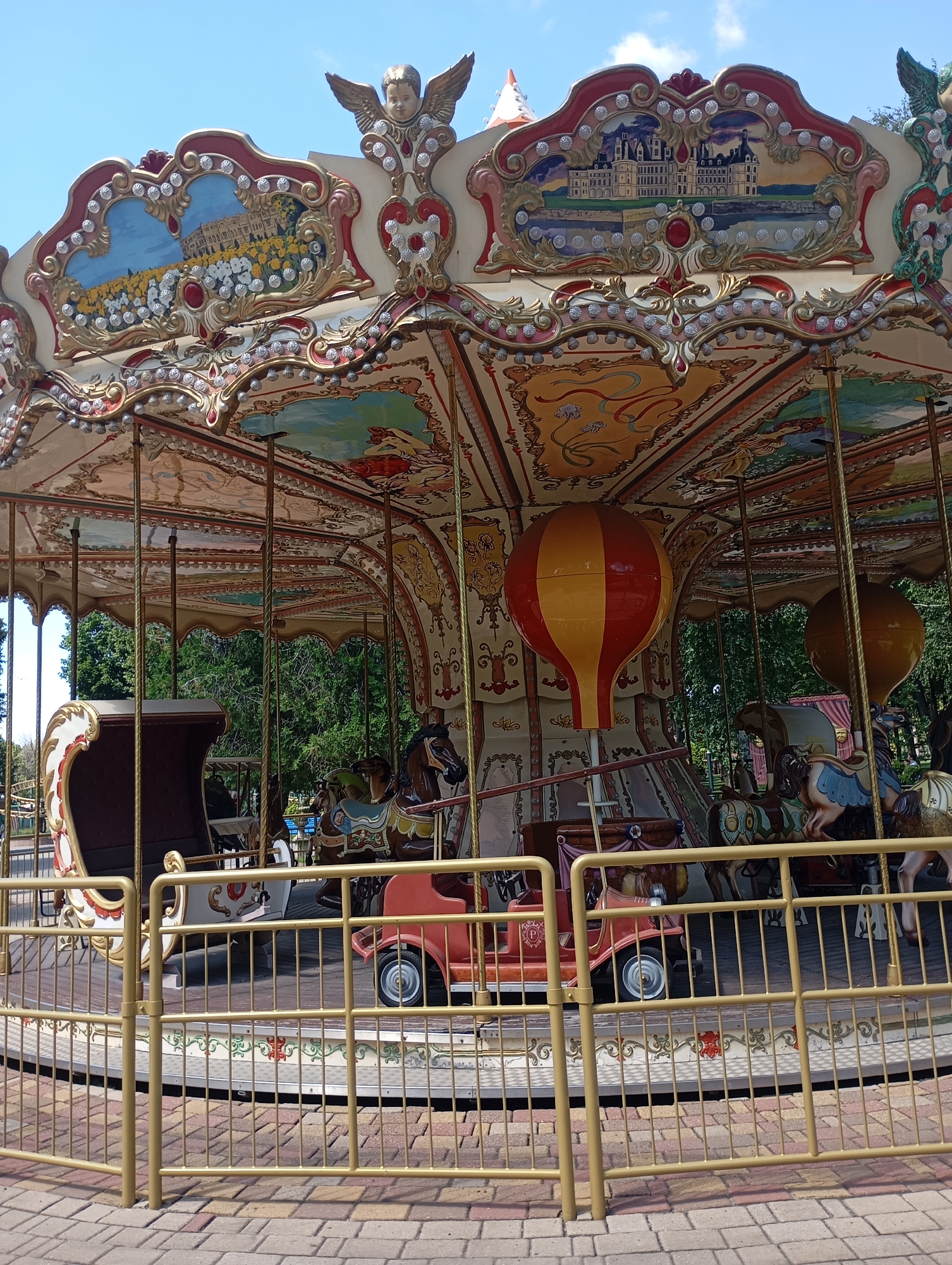 The image shows a carousel in a park on a sunny day. The carousel has a colorful roof with painted scenes and decorations, including an angel at the top. There are various figures on the carousel, such as horses and a vintage car. A hot air balloon decoration is also visible. The carousel is surrounded by a yellow metal fence, and beyond that, there are trees and a paved walkway. The location is identified as Kharkiv, Ukraine.