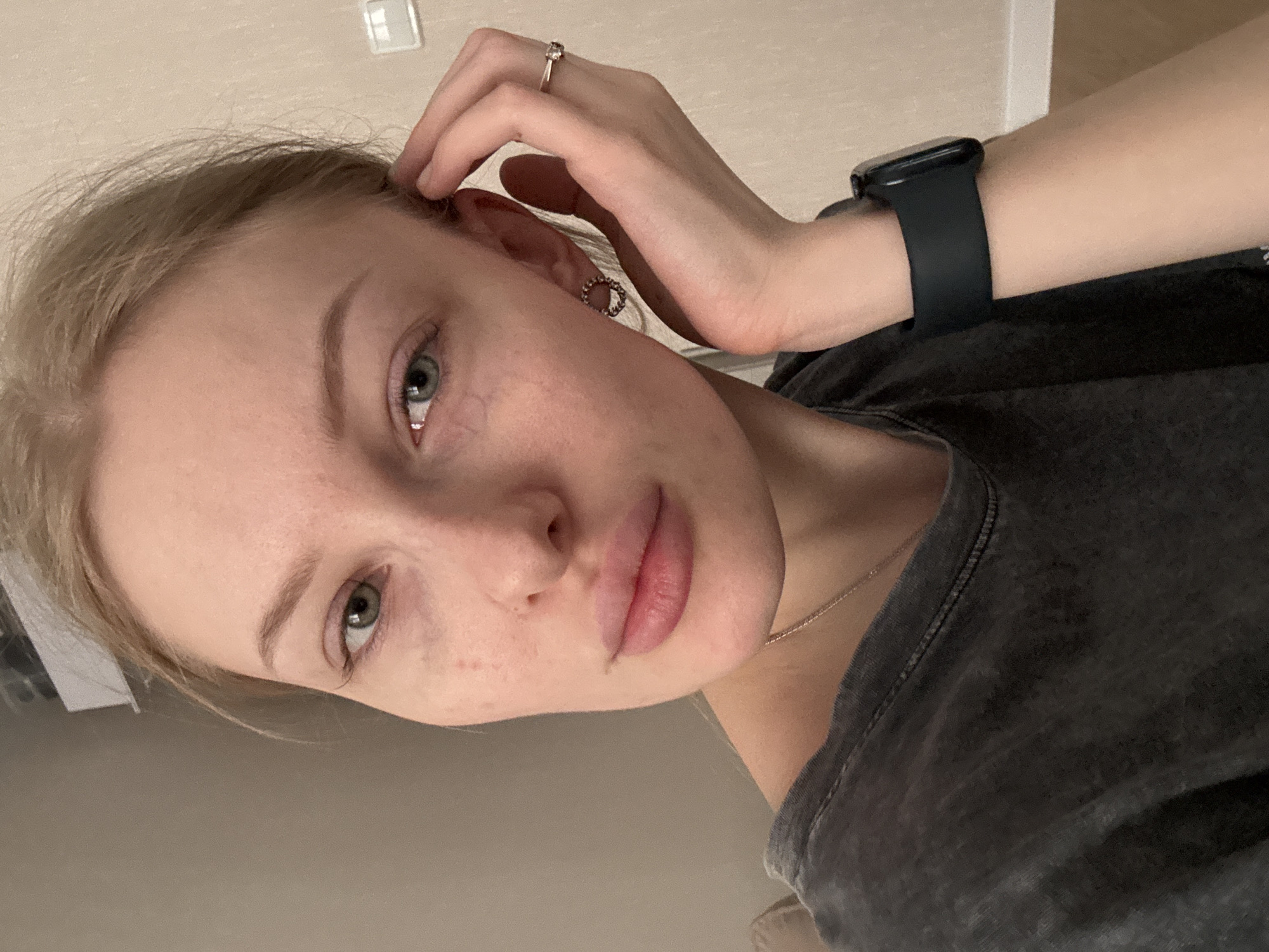 A young, fair-skinned woman with blonde hair and light-colored eyes is depicted in a close-up shot. She is wearing a dark grey t-shirt and a black smartwatch on her left wrist. A ring is visible on a finger of her left hand, which is raised and positioned near her ear. She has minimal visible makeup. The background is a neutral, light-colored wall with a light switch. There is no indication of Perm, Russia in the image. The scene is indoors.