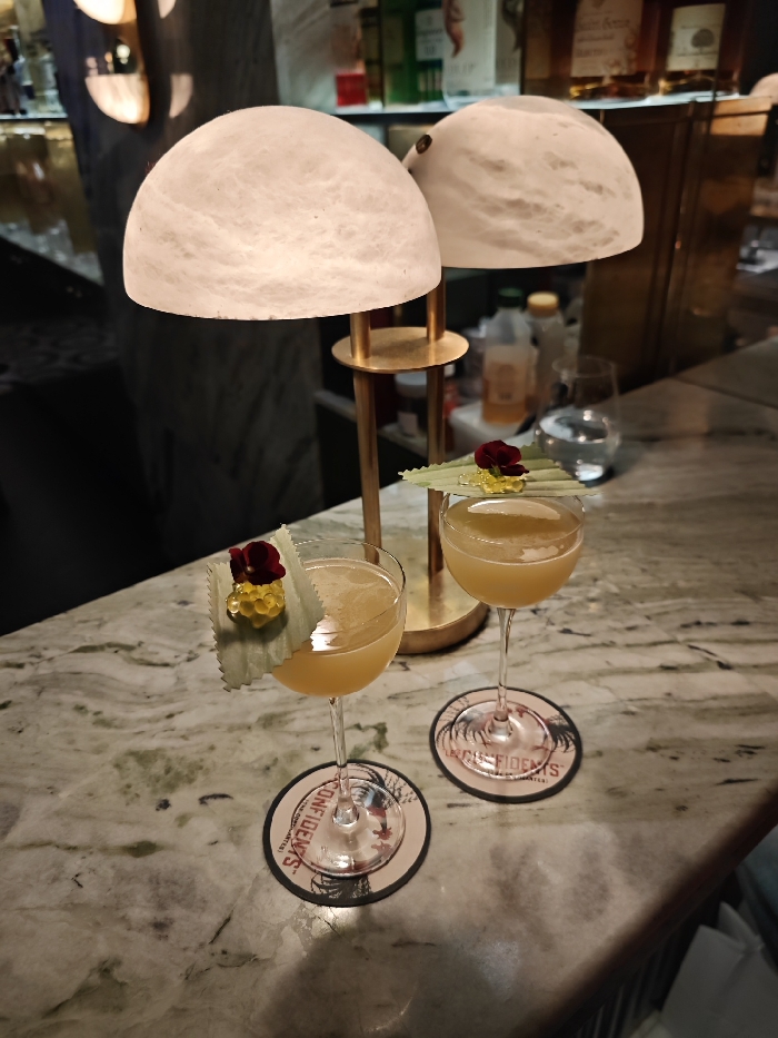 Two cocktails sit on a marble bar. Each is garnished with a leaf, caviar-like spheres, and a small flower. Both cocktails rest on coasters with "CONFIDENTS" printed on them. Behind the drinks are two table lamps with white, semi-spherical shades. The lamps have gold-colored bases. In the background are various bottles and glassware on a bar shelf.