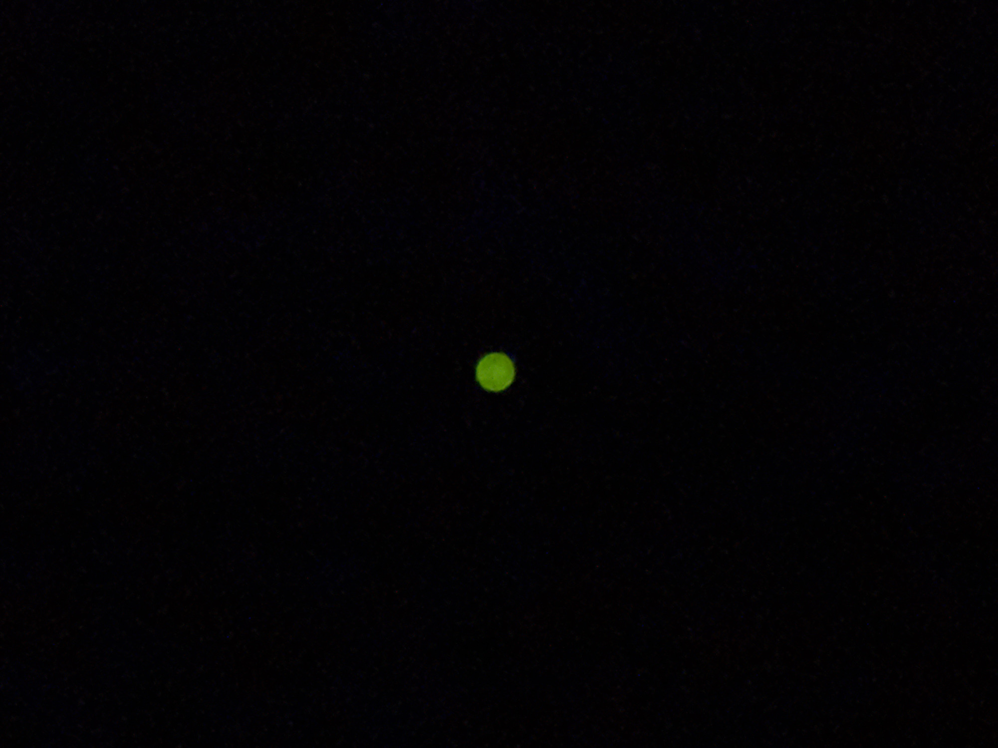 The image is a close-up shot of a single, small, green circular light against a black background. The light appears to be a discrete point source, possibly an LED or a small illuminated object. The background is uniformly dark, with no discernible features or details. The image does not provide any information or context about its location in Burgas, Bulgaria, or any other specific details about people, subjects, objects, actions, interactions, or the environment.