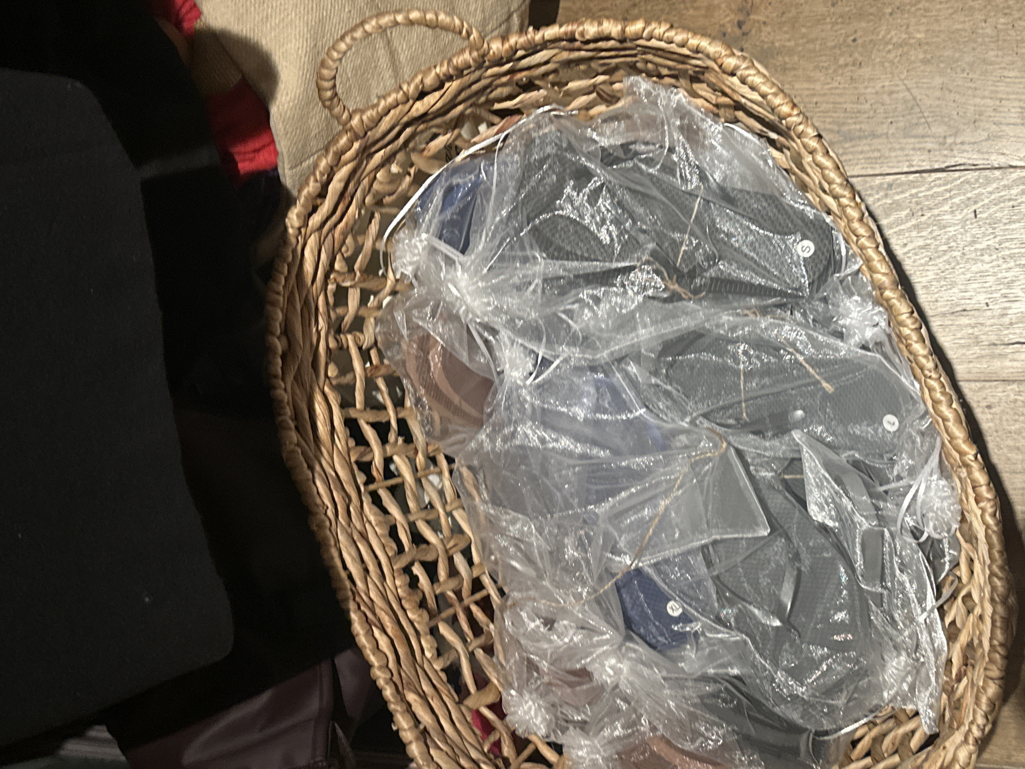 The image shows a woven basket filled with items covered in clear plastic packaging. The basket is sitting on a wooden floor. To the left of the basket is a dark-colored object that appears to be soft, possibly a piece of furniture. The scene's location and any people present cannot be determined from the image. It is impossible to determine if the image was taken in London, United Kingdom.
