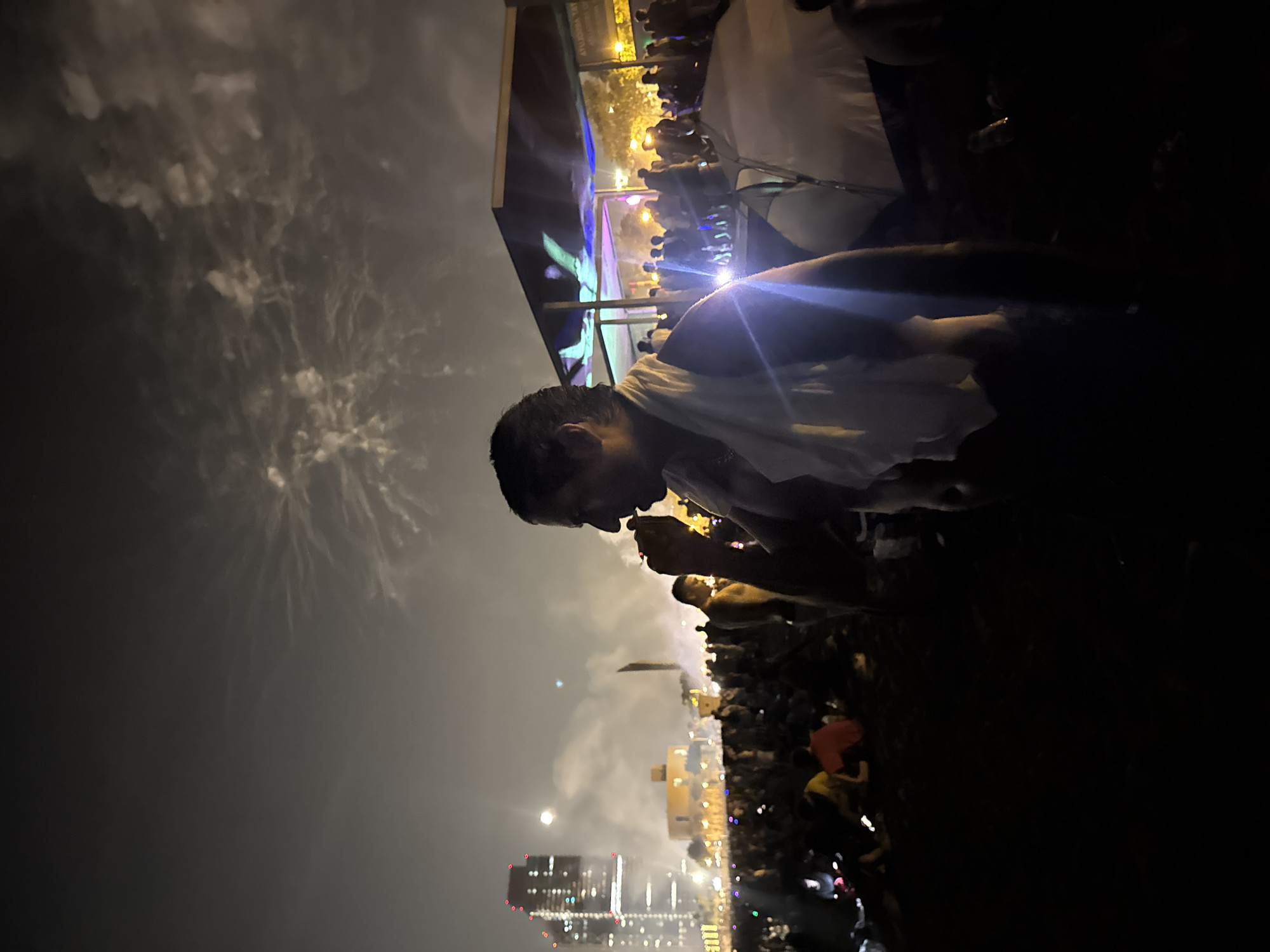 The image depicts a nighttime scene with fireworks in the sky over Barcelona, Spain. In the foreground, a man is silhouetted against the brighter background, appearing to hold something to his mouth. He is wearing a white towel draped over his shoulders. A crowd of people is gathered, many sitting on the ground. In the mid-ground, there are tents and a covered structure with colorful lights. In the background, city buildings are visible, including tall, illuminated structures. The overall atmosphere suggests a public gathering or celebration.