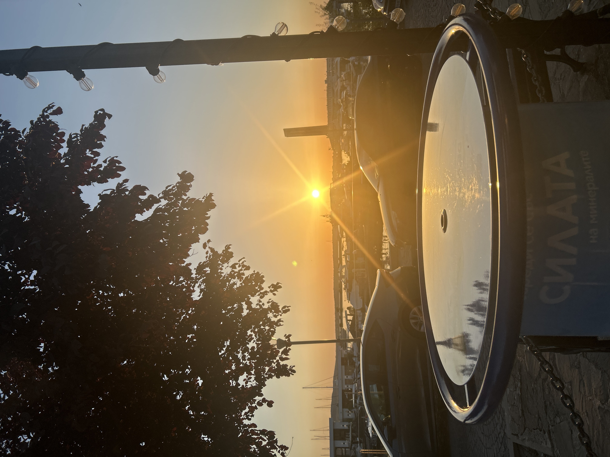 The image depicts a sunset scene in Sozopol, Bulgaria, likely near the waterfront or a marina. In the foreground, a round table with a reflective surface sits on a cobblestone ground, partially obscured by a dark-leafed tree branch hanging from the upper left. The table has a blue rim and a light blue base with Cyrillic text that reads "СИЛАТА на минералите," translating to "The Power of Minerals." A metal chain fence runs along the edge of the table.
Beyond the table, two dark-colored cars are parked on a paved area. The car on the left is a sedan, and the car on the right is also a sedan or hatchback. In the background, a marina is visible, filled with numerous docked sailboats and yachts. Buildings line the waterfront, and beyond them, the horizon is bathed in the warm glow of the setting sun, which is positioned slightly to the left of the center of the frame, casting long rays. The sky transitions from a bright orange near the horizon to a clear blue higher up.
To the right of the frame, a tall, dark pole is adorned with a string of clear, Edison-style light bulbs, some of which are illuminated. The light from the sunset reflects off the surface of the table and the cars. The overall impression is a serene evening ambiance in a coastal town. There are no people clearly visible in the image.