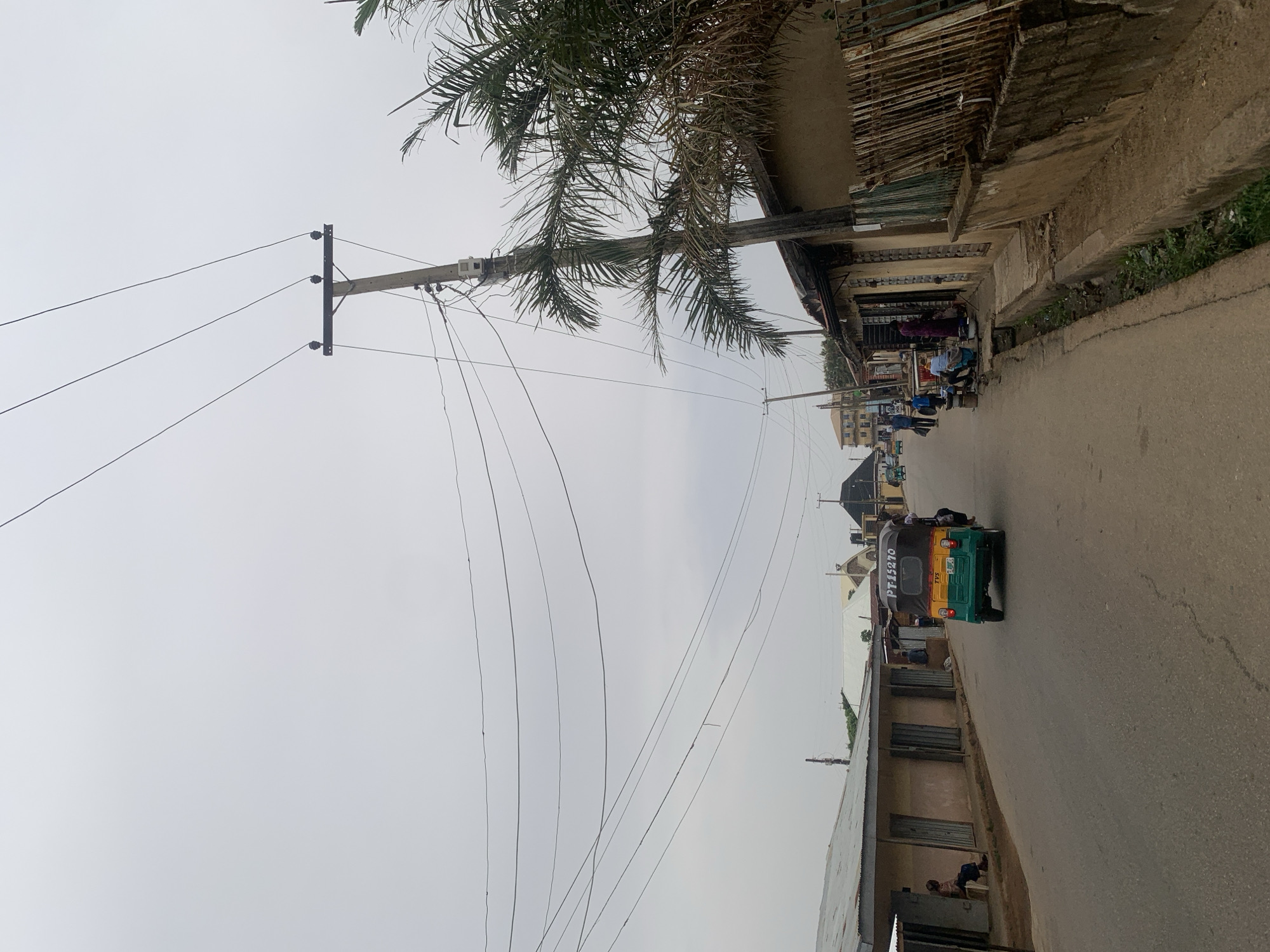 The image depicts a street scene in Jos, Nigeria, observed from a slightly elevated position. A paved road stretches into the distance, where buildings are visible. An auto-rickshaw, painted green, yellow, and black, is moving along the road. The sides of the street are lined with buildings, some appearing to be shops or businesses. Utility poles with numerous electrical wires are prominent on the left side of the frame. A palm tree is also visible, its fronds extending over the street. People are seen in the distance, near the buildings on the side of the road. The sky is overcast.