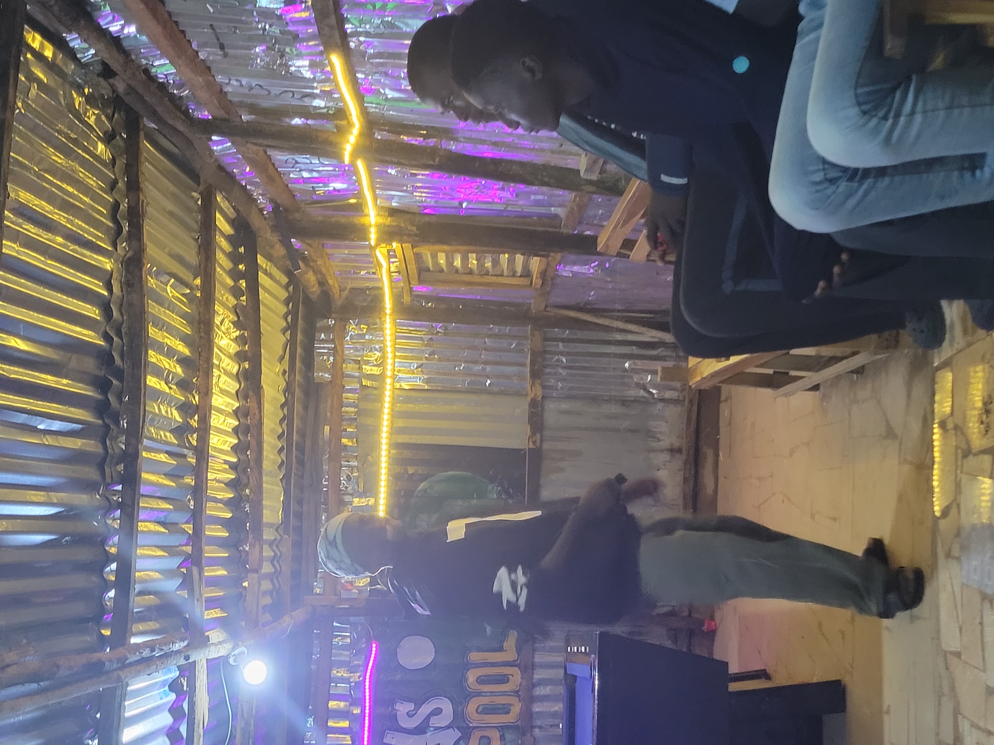 The image shows an indoor setting, possibly a bar or entertainment venue, with a corrugated metal roof and walls. The space is dimly lit with string lights, some yellow and some purple. In the foreground, a person in a dark jacket and light-colored pants is standing and looking towards the right of the frame. To the right, three individuals are seated on a bench, facing away from the camera. One person is wearing a dark hoodie and light blue jeans, another is wearing a dark jacket, and the third person's attire is less visible. A pool table is partially visible on the left side of the image. The floor is tiled. There is a sign with the letters "S POOL" illuminated in neon lights. The setting appears to be informal and possibly a local establishment in Durimi, Nigeria.