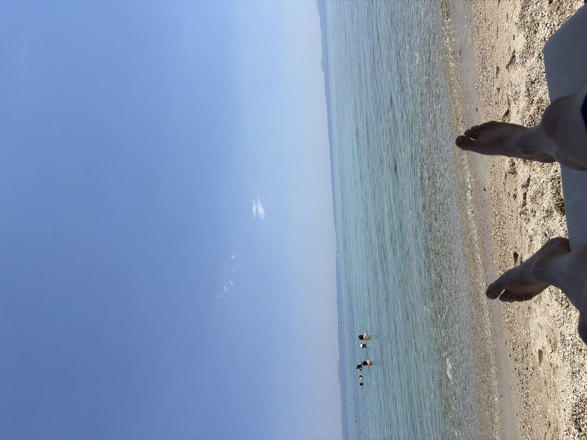 The image depicts a serene beach scene with crystal-clear, shallow turquoise water lapping onto a sandy shore. The perspective is from someone reclining on the beach, with their bare feet in the foreground, lightly dusted with sand. In the distance, several people are visible wading and swimming in the calm sea. The sky is a vast, clear blue, with a single, wispy cloud adding a touch of texture. The horizon shows a faint outline of land, suggesting a coastline or islands in the distance. The bright, clear sky indicates it's a sunny day, likely in the late morning or early afternoon. The overall atmosphere is one of relaxation and enjoyment of a beautiful day at the seaside. The location is identified as Kassandra Municipal Unit, Greece, known for its picturesque beaches and clear waters. No text is visible in the image.