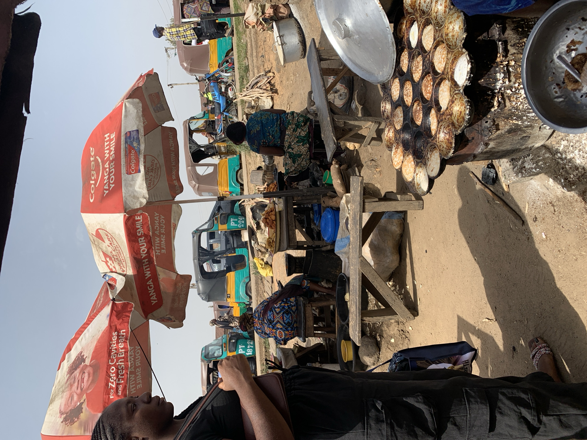 The image captures a vibrant street scene, likely in Rafin Pa, Nigeria, judging from the signs in the image. Dominating the foreground are Colgate-branded umbrellas with "YANGA WITH YOUR SMILE" visible on them. A woman in black clothing and braided hair is standing or walking on the left side of the frame.
Further into the scene, two women are seated on low wooden stools, seemingly engaged in preparing and selling food. One appears to be cooking in a large, well-used pan with multiple circular indentations containing batter or food. Next to the cooking setup is a collection of pots, firewood, and other cooking essentials.
Behind the food preparation area, a line of auto rickshaws (tuk-tuks) in shades of yellow, green, and silver are parked, suggesting a busy transportation hub. Several people are visible in and around the tuk-tuks, implying activity and movement. The environment is a dusty, open space, which indicates an urban or semi-urban setting.