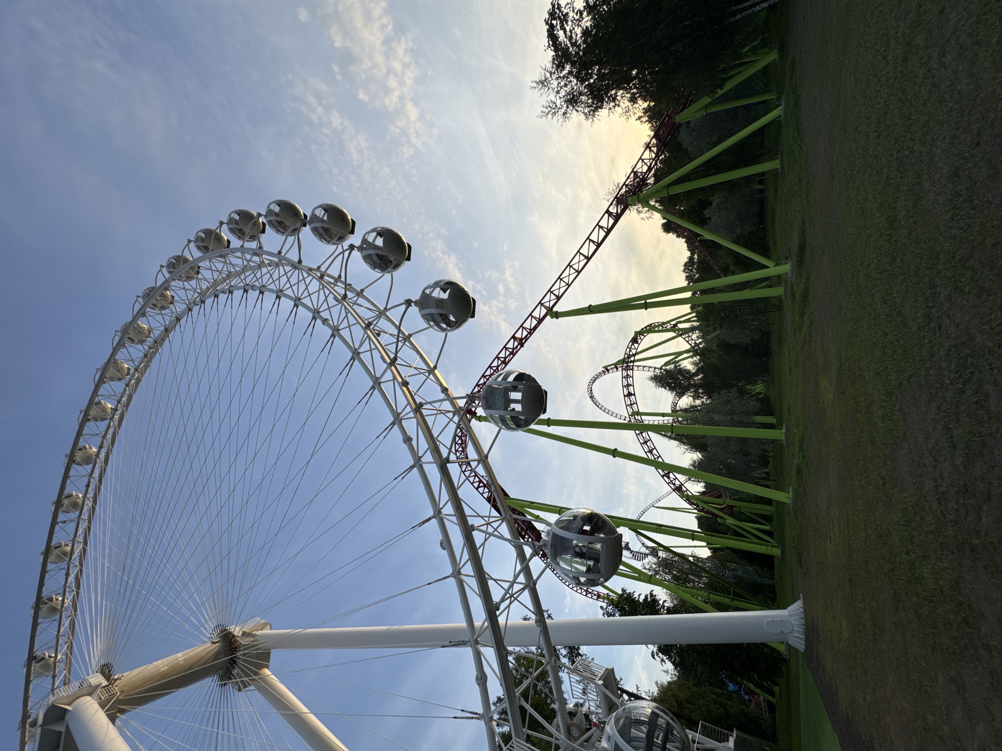 The image shows a scene at an amusement park in Saint Petersburg, Russia, featuring a Ferris wheel and a roller coaster. The Ferris wheel, white in color, has a large circular structure with numerous gondolas suspended from it. The roller coaster track, predominantly red and supported by lime-green poles, winds through the scene, with loops and drops visible. The environment includes a grassy foreground with some patches of dirt, and a backdrop of trees. The sky is a clear blue with some wispy clouds, and the lighting suggests it is either early morning or late afternoon, as there is a soft, warm glow on the horizon. Several gondolas on the Ferris wheel and sections of the roller coaster track are clearly visible. The image is taken from a low angle, looking upwards towards the attractions.