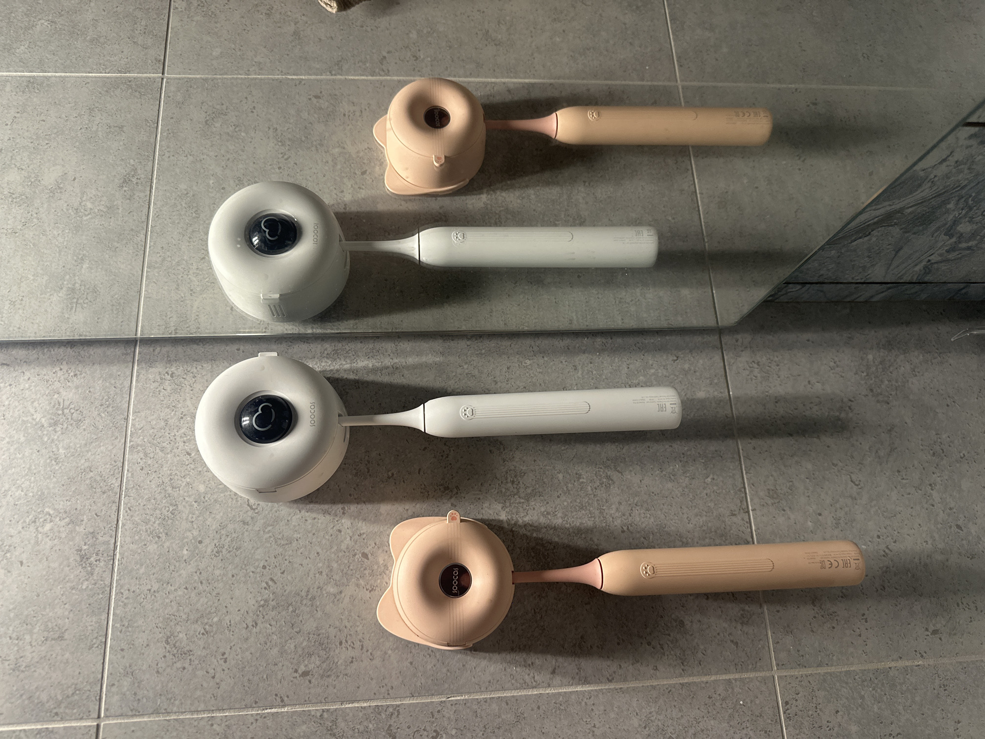 Two electric toothbrushes are mounted on a tiled wall, reflected in a mirror. The toothbrush on the left has a peach-colored handle and a pink cat-shaped holder. The toothbrush on the right has a white handle and a white circular holder. Both holders have a black circular lens with a heart-shaped reflection visible inside. The wall is made of large, gray, square tiles. The floor, partially visible in the mirror, appears to be a dark, marbled surface. The scene is clean and well-lit, suggesting a domestic bathroom. There is no indication of the location being in Moscow, Russia.