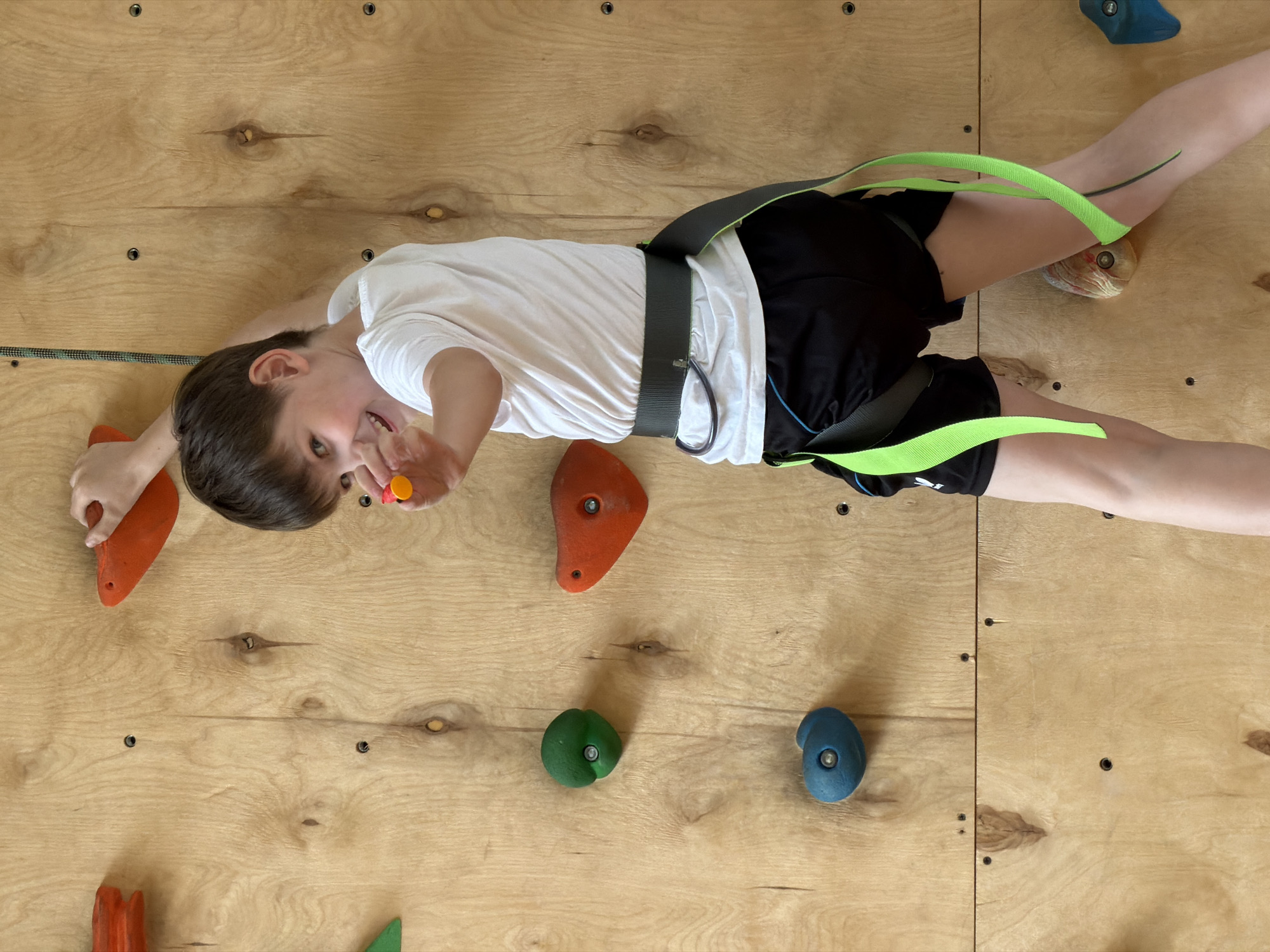 A boy is climbing a climbing wall. The boy is wearing a white t-shirt, black shorts, and a climbing harness. He is holding onto two orange climbing holds with his hands, and his feet are spread apart, with one foot on a light-colored climbing hold. He is smiling and looking at the camera, and he is holding a small, yellow object in his right hand. The climbing wall is made of wood and has a variety of climbing holds attached to it. The city of Izhevsk, Russia is not evident in this image.