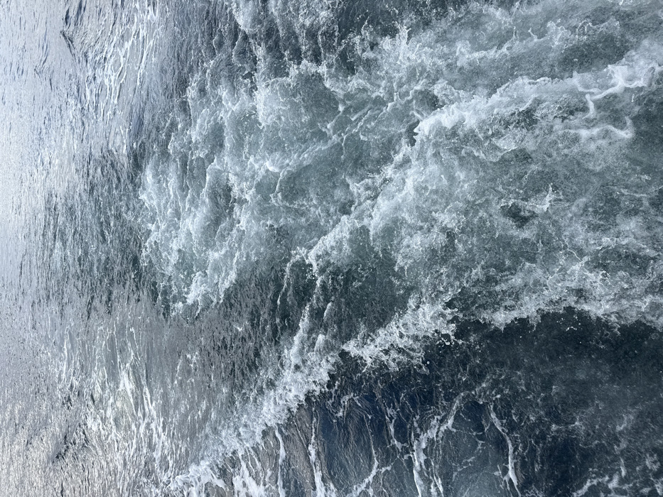 The image displays a turbulent boat wake across a body of dark blue-grey water, indicative of the marine environment near Ushuaia, Argentina. A broad, frothy white trail of disturbed water dominates the lower and central portions of the frame, tapering into less defined ripples towards the upper area. The water exhibits significant churning and aeration, characterized by various shades of deep blue, charcoal grey, and bright white foam. No landmasses, sky, vessels, individuals, or other distinct objects are visible within the frame. The scene is solely comprised of the agitated surface of the water, documenting the immediate aftermath of a vessel's passage through open waters.
