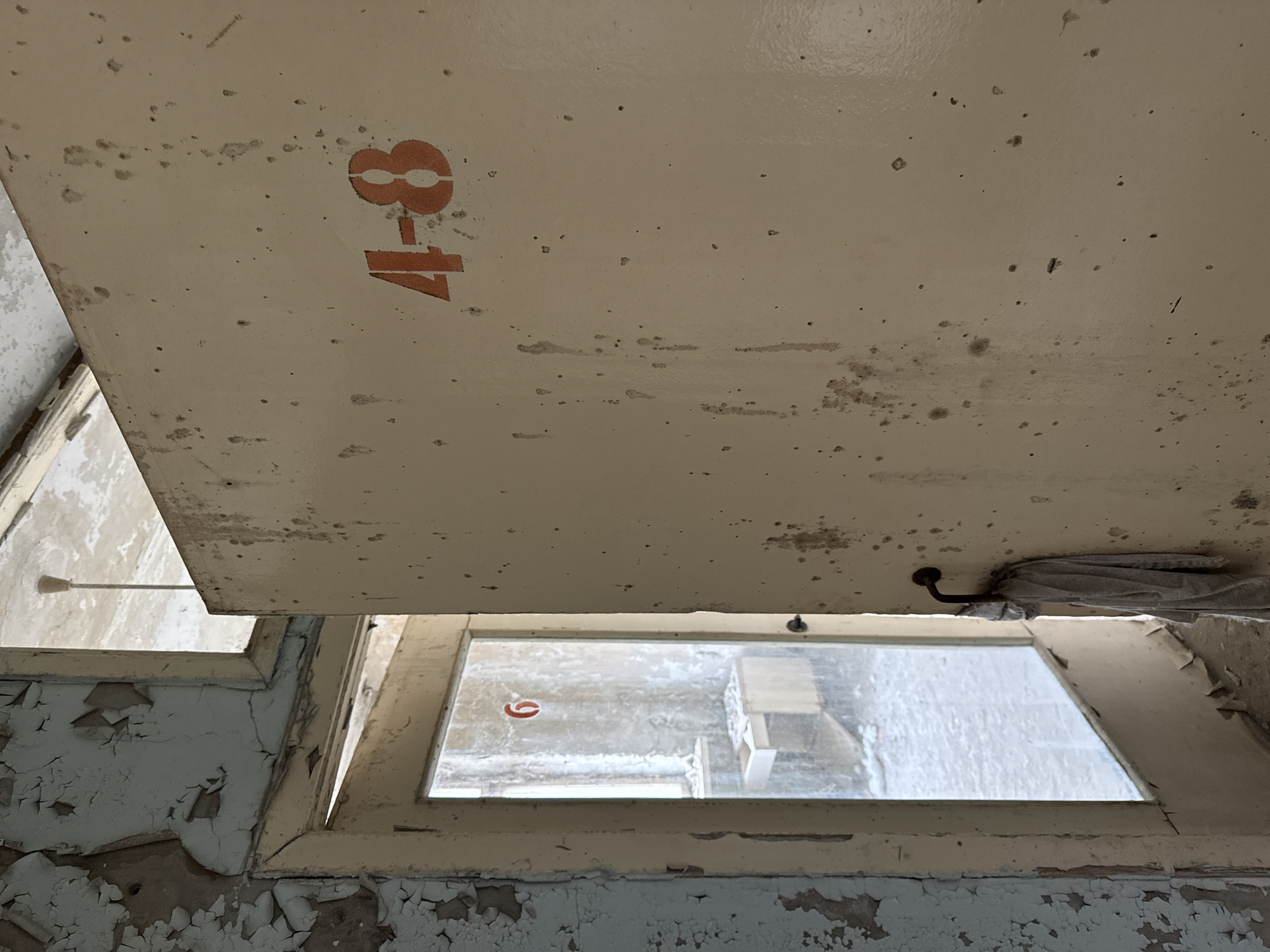 The image depicts a decaying interior, likely in a building located in Prypiat, Ukraine, based on the state of disrepair and architectural style. The primary focus is on a cream-colored ceiling or overhead panel with the number "8-17" stenciled in reddish-orange. The panel is heavily mottled with stains and signs of deterioration.
Below the panel is a window frame with peeling paint and a view of a blustery, snowy and possibly abandoned exterior. Through one of the panes of glass a red circle is visible, possibly a sticker. The wall surrounding the window shows extensive paint peeling, indicating significant neglect. A hook with an indiscernible object hanging from it is visible on the ceiling above the window. The image presents a picture of abandonment, decay, and dereliction.