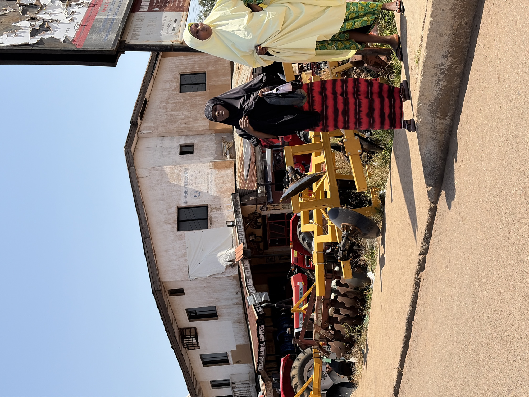 Two women stand on a sidewalk in front of a building that appears to be a business. The woman on the left wears a black hijab and a red and black striped skirt. The woman on the right wears a yellow hijab and a green and yellow patterned skirt. Behind them, the building has several windows and a sign that reads "CHEKNUS GLOBAL SERVICES". Several tractors and agricultural equipment are parked in front of the building. The scene appears to be in a city, with a road in the foreground and buildings and sky in the background. The image is dated to November 2023. The location is identified as Gura Top, Nigeria.