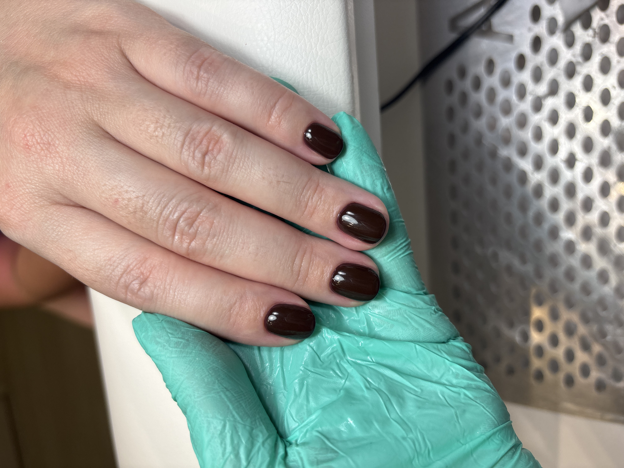 A close-up shot displays a fair-skinned hand with short, dark brown nails resting on a green gloved hand. The hand is positioned on a white surface, and the background includes a metal object with a perforated pattern. The setting appears to be indoors.