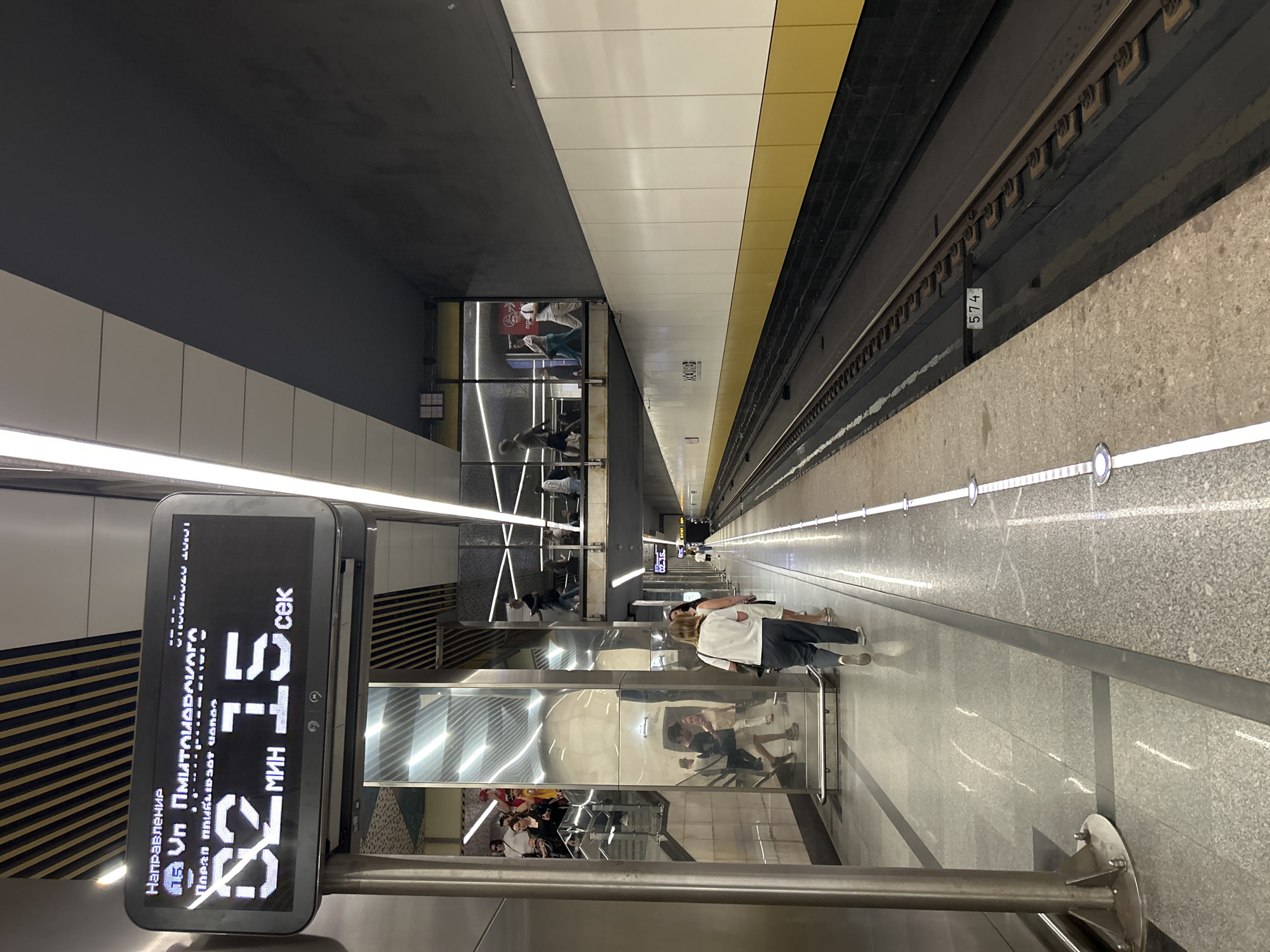 The image captures a scene at a Moscow Metro station. The central focus is a digital display showing the time to the next train: "02:15" (2 minutes and 15 seconds). The display also indicates the direction of travel as "ул. Дмитриевского" (Dmitrievskogo Street) and "Парк Сокольники" (Sokolniki Park). Above this, a timestamp reads "07.08.2020 15:01". The station platform is modern, with polished granite floors and sleek metallic columns. Several people are visible on the platform, some walking and others waiting. In the background, an elevated walkway or mezzanine level is visible, with more people moving around. The train tracks run along the platform. The station walls are decorated with white panels and a yellow band. The overall impression is that of a clean, well-lit, and contemporary urban transit environment.
