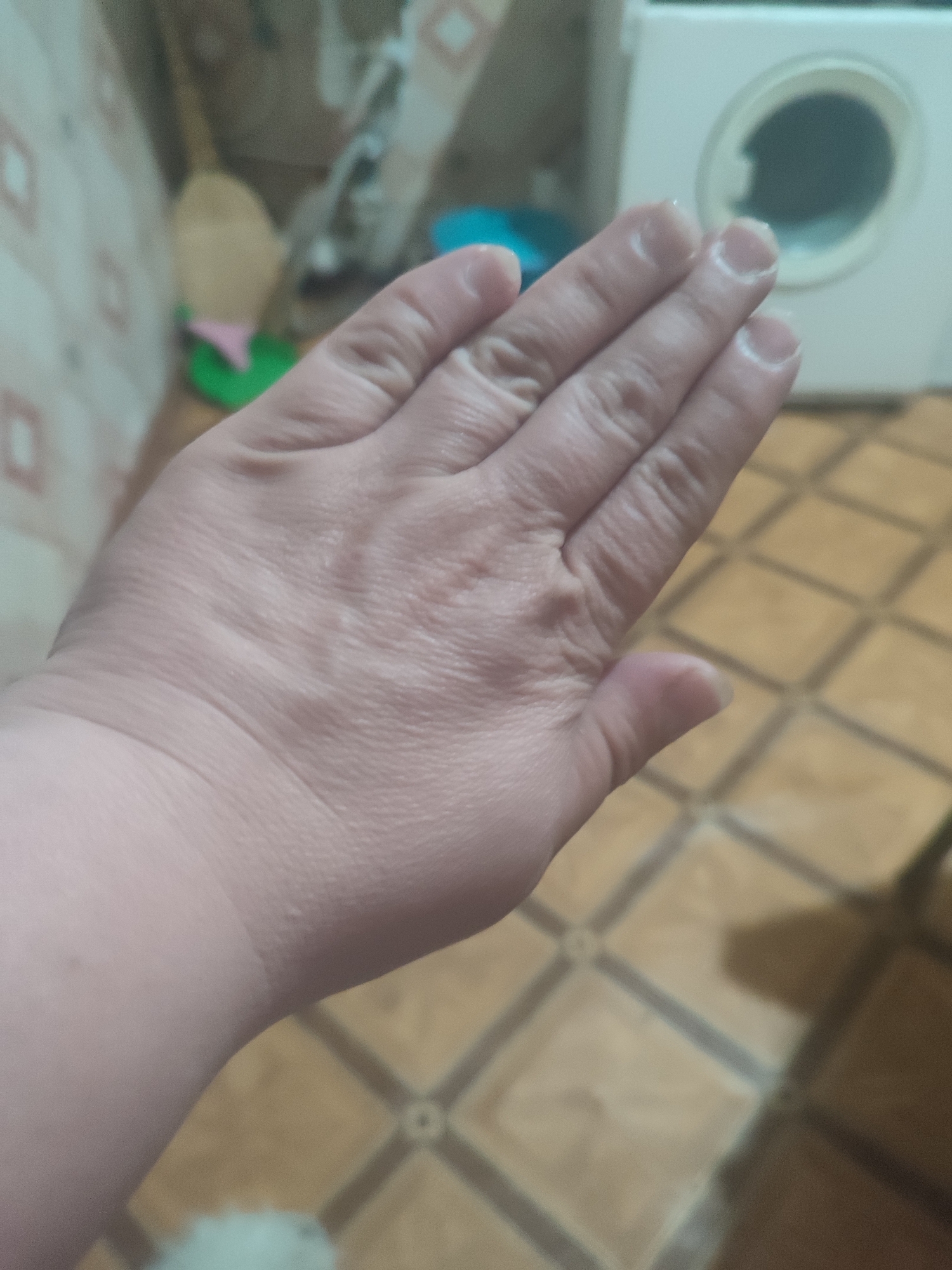 An indoor, eye-level, close-up shot shows a left hand, palm up, with fingers extended. The hand is lightly wrinkled and appears to be facing a blurred background. The background includes a portion of a white appliance with a circular opening, possibly a washing machine, and patterned floor tiles. No specific location within Chernivtsi, Ukraine, can be determined from the image.