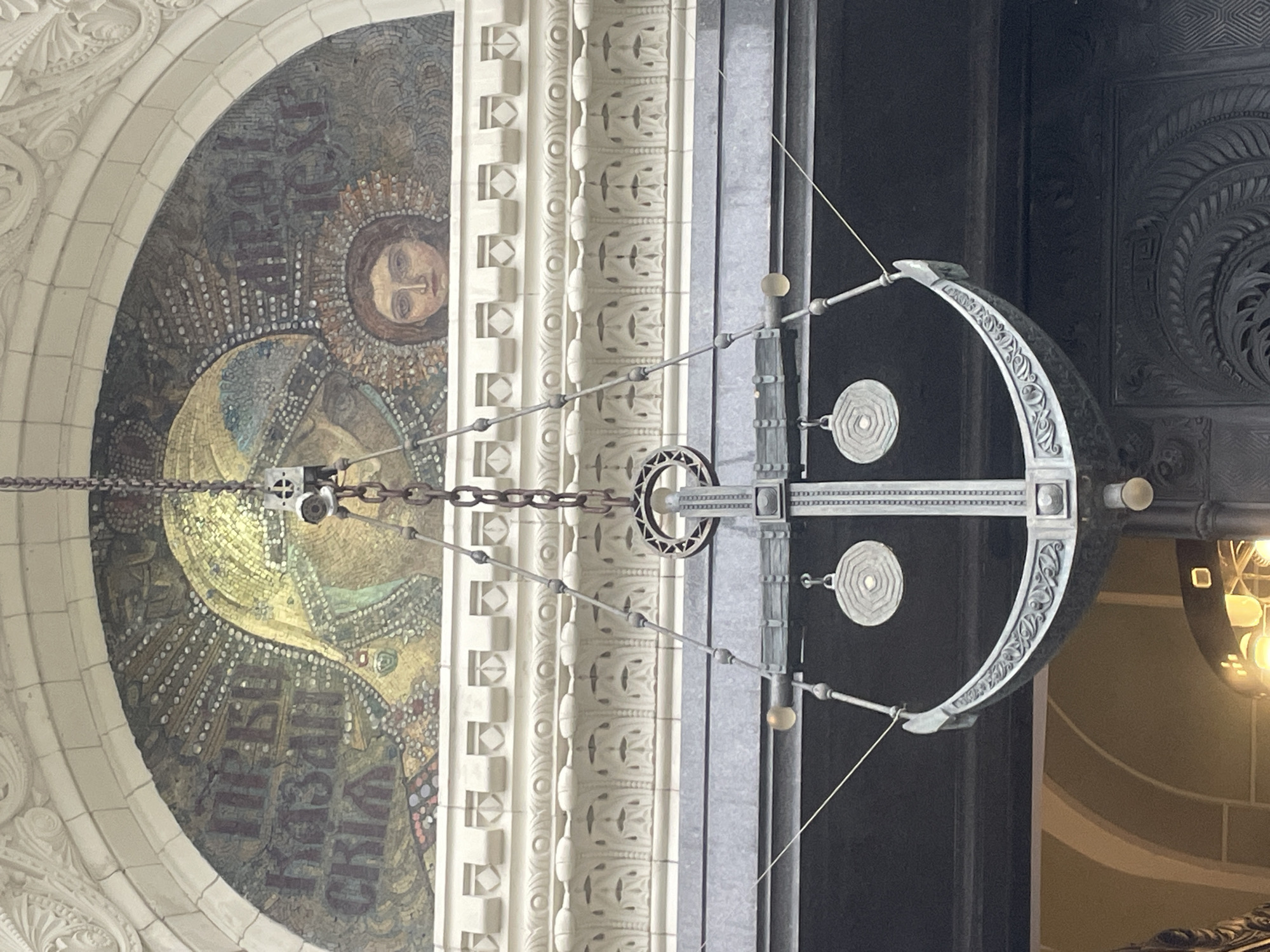 The image shows a mosaic of the Virgin Mary and Christ Child above an ornate chandelier that is shaped like a smiley face. The mosaic is located on the facade of the Naval Cathedral of Saint Nicholas in Kronstadt, Russia. The mosaic is made of small, colorful tiles and depicts the Virgin Mary with her hand extended, holding the Christ Child. The chandelier is made of dark metal and has two circular discs that resemble eyes, and a curved bar that forms a smile. The chandelier is hanging from a chain and is illuminated by several light bulbs. The background is a dark opening, possibly an entrance to a building. The overall scene suggests a juxtaposition of religious art and decorative architecture.