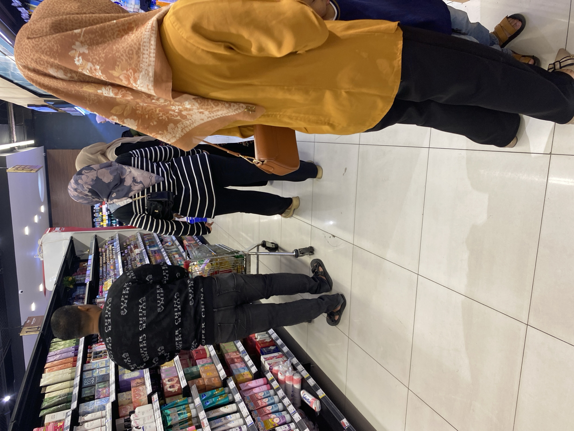 In a brightly lit supermarket aisle in Banda Aceh, Indonesia, a man wearing a black sweatshirt with "SUPER CHINA" printed repeatedly in white, and dark wash jeans, stands near a shopping cart. He is facing away from the camera, looking towards shelves stocked with various products, including tubes of lotion and boxed items. To his right, a woman in a striped long-sleeved shirt and black pants, wearing a patterned headscarf, walks ahead. Further to the right, another woman in a mustard-yellow top and black pants, with a floral patterned headscarf, is seen from behind. She carries a brown handbag. The floor is tiled in a light, neutral color with visible grout lines.
