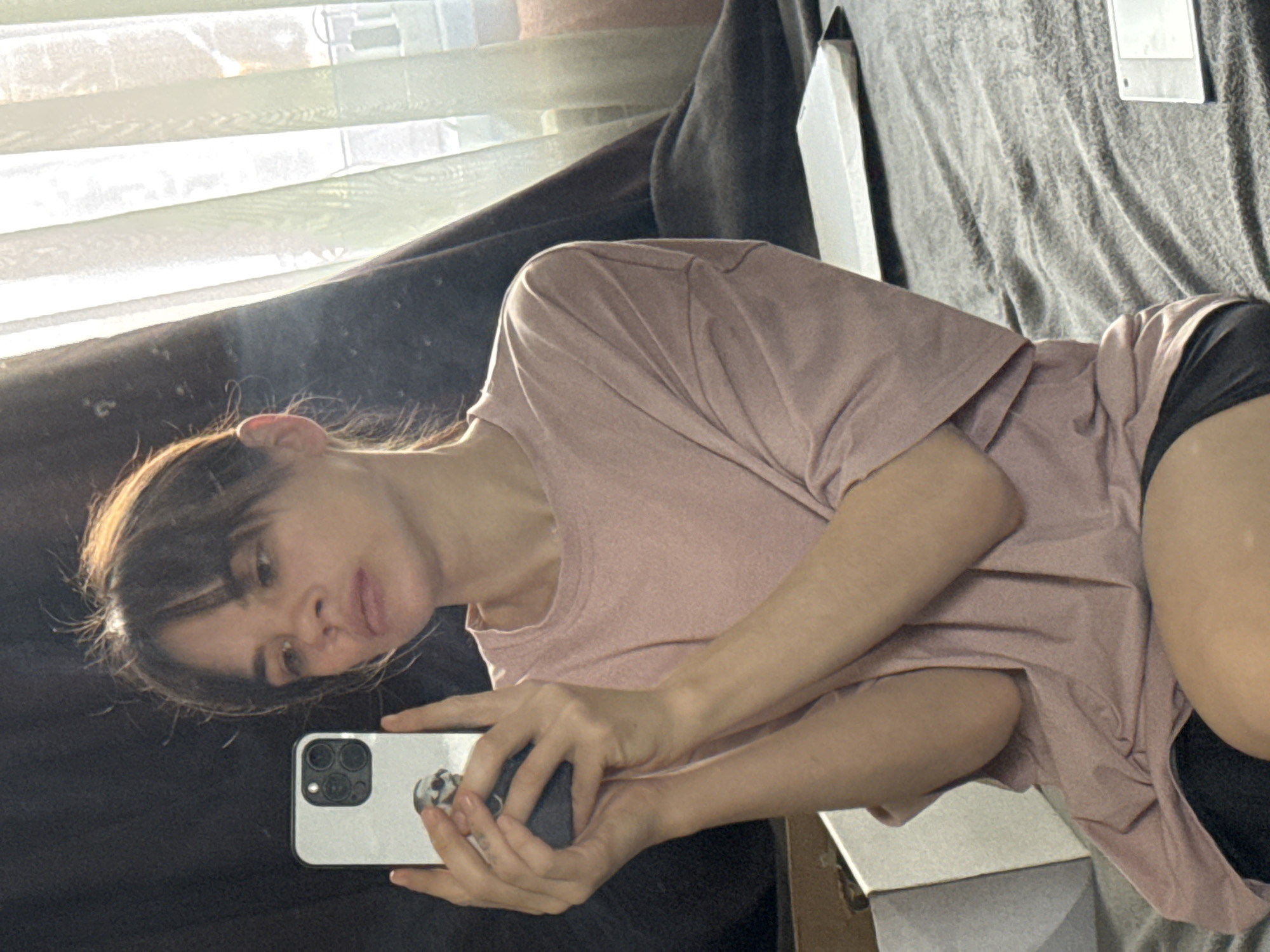 A woman with dark hair and bangs is holding a white smartphone with multiple camera lenses in her hands. She is wearing a pink t-shirt and black shorts. The background includes a window with blinds, a dark curtain, and a gray couch. There is also a white object, possibly a piece of furniture, visible near her arm. The location is likely indoors. There is no indication that this is located in Rostov-on-Don, Russia, as there are no identifiable landmarks to specify the location.