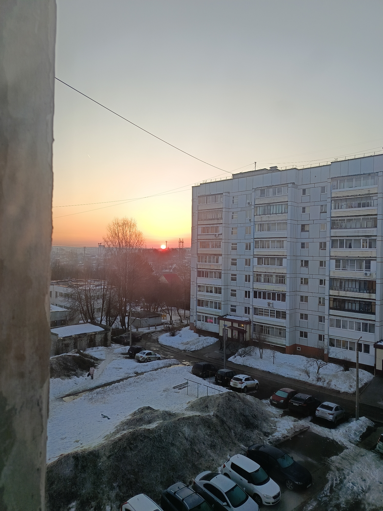 An elevated, outward-facing view captures an urban landscape in Chaikovsky, Russia, during winter, likely at sunrise or sunset given the low sun angle and warm light on the horizon.
The immediate left foreground is dominated by a vertical, light-grey textured concrete or plaster architectural element. A thin, dark cable traverses the upper sky from this element towards the right.
The sky transitions from a pale blue-grey at the zenith to a vibrant orange-red at the horizon, where the sun is visible as a distinct red-orange sphere, partially obscured by bare trees and distant industrial structures including power lines. A faint, thin contrail is present in the upper-left sky.
The midground features a prominent multi-story (approximately 9-10 floors), light-grey residential apartment building with a grid-like facade, numerous windows, and enclosed balconies. The ground surrounding the building and in the central midground is extensively covered in snow, forming irregular mounds, some appearing dirty or disturbed. Darker asphalt pathways and parking areas are visible where snow has been cleared or melted.
Numerous passenger vehicles are parked throughout the scene, including white, black, silver, and red cars, some partially covered in snow.
One individual is observed walking on a cleared path through the snow in the lower-middle left, wearing a light-colored, possibly pink or light brown, coat. Another figure is faintly visible near the ground-level entrance of the large apartment building.
In the background, beyond the immediate residential area, bare deciduous trees are visible, along with smaller, single-story structures and a hazy expanse of a distant city or industrial area under the low sun.