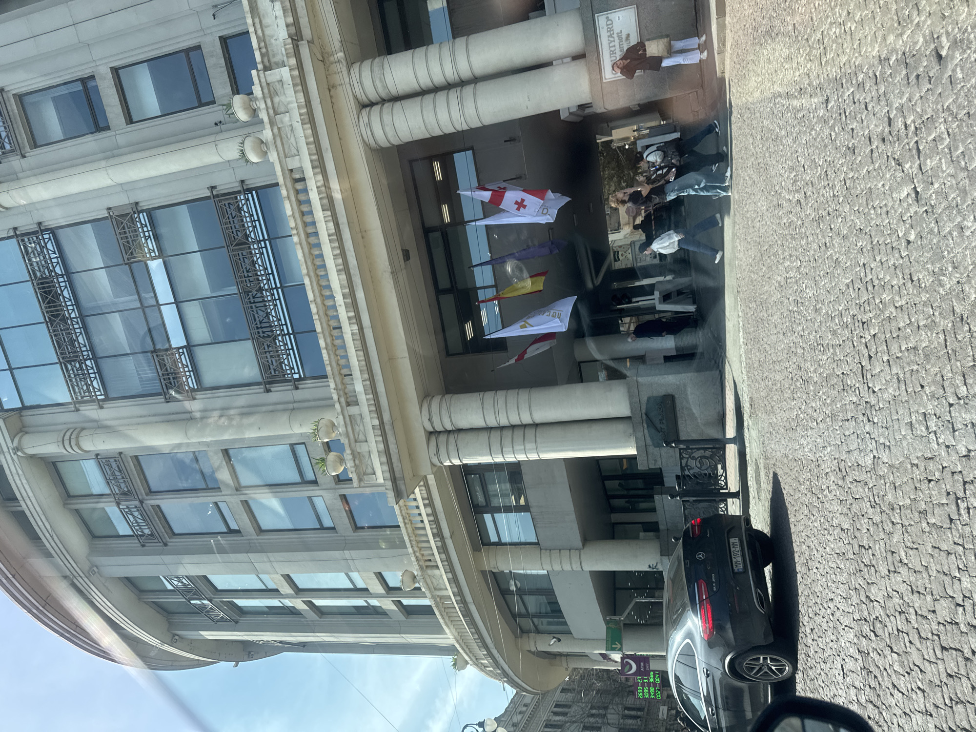 The image shows a street scene in Tbilisi, Georgia, specifically the Courtyard Marriott Hotel, identified by the signage. The hotel facade is visible with classical architectural elements like columns and ornate window detailing. Several flags are hanging, potentially representing different countries or organizations, including what appears to be the flag of Georgia with its distinctive red crosses. People are seen near the hotel entrance. A black Mercedes GLC 250 is parked on the cobblestone street, bearing the license plate MM-192-MM. Street signage with the text ATM and numbers is visible in the lower left, and "EMT PLAZA" is written on a sign above the hotel entrance. The image seems to be taken from inside a vehicle.