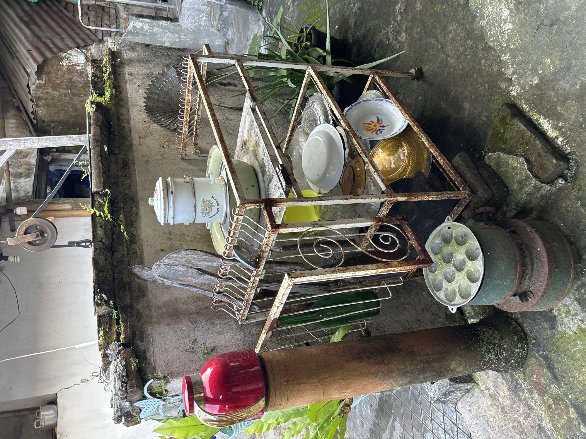 In Banguntapan, Indonesia, an outdoor scene depicts a rustic storage rack laden with various kitchenware. A red teapot with gold accents stands prominently on a terracotta pipe, juxtaposed with lush green foliage. The rack itself, weathered and rusty, holds stacked metal containers, bowls, and plates. Below, a circular mold sits atop a rusted stove, hinting at past culinary activities. The surroundings feature a concrete floor, a weathered wall, and distant glimpses of a corrugated iron roof, all indicative of an aged, utilitarian space.