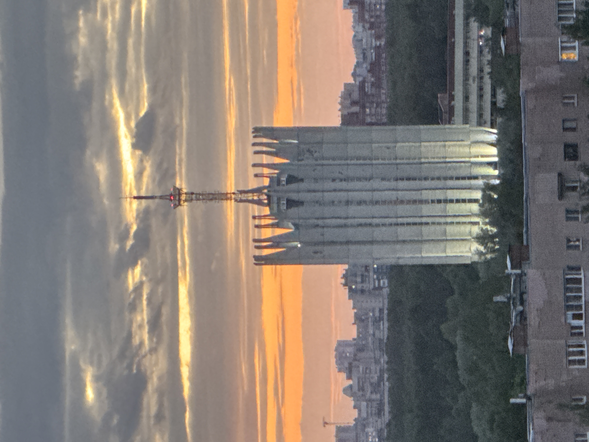 A vertical shot shows an urban cityscape during sunset. In the background, there's a striking sunset with a blend of golden and grey clouds, creating an interesting color contrast. A communications tower is visible against this colorful backdrop. To the right of the tower, there's a multi-level building with a unique architectural design, featuring protruding horizontal sections. The cityscape with various buildings and greenery extends from the right side towards the bottom. The scene has a tranquil, urban feel, capturing the beauty of a sunset in a city environment.