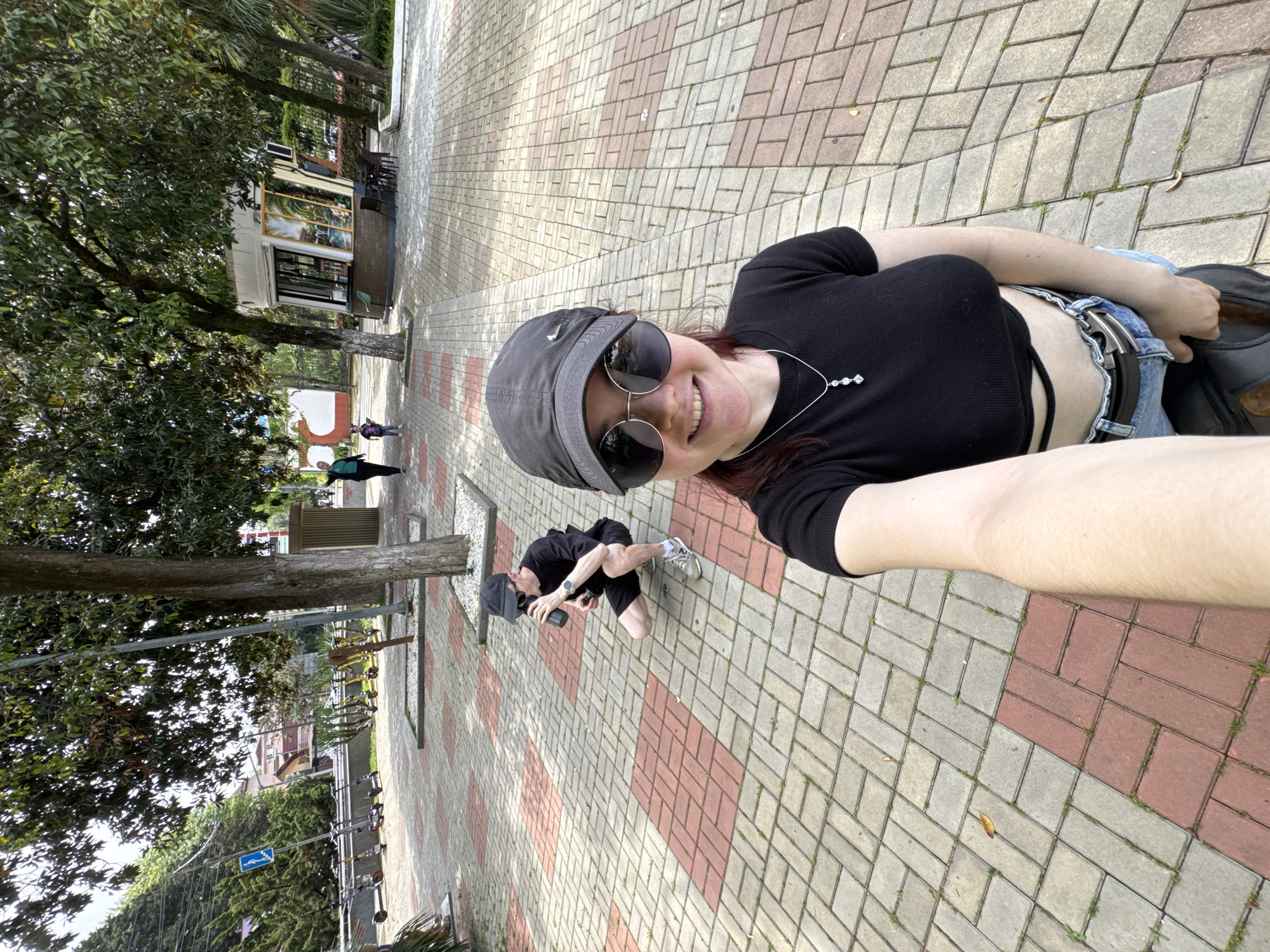 A medium shot shows a woman taking a selfie on a pedestrian walkway. The walkway is paved with rectangular bricks, arranged in alternating rows of red and grey. The woman wears a black crop top, jeans, sunglasses, and a baseball cap. She is smiling at the camera. In the background, a man wearing similar attire is squatting and appears to be taking a photo. The setting appears to be the city of Sochi, Russia, possibly a park or public space, given the trees, buildings, and rental scooters.