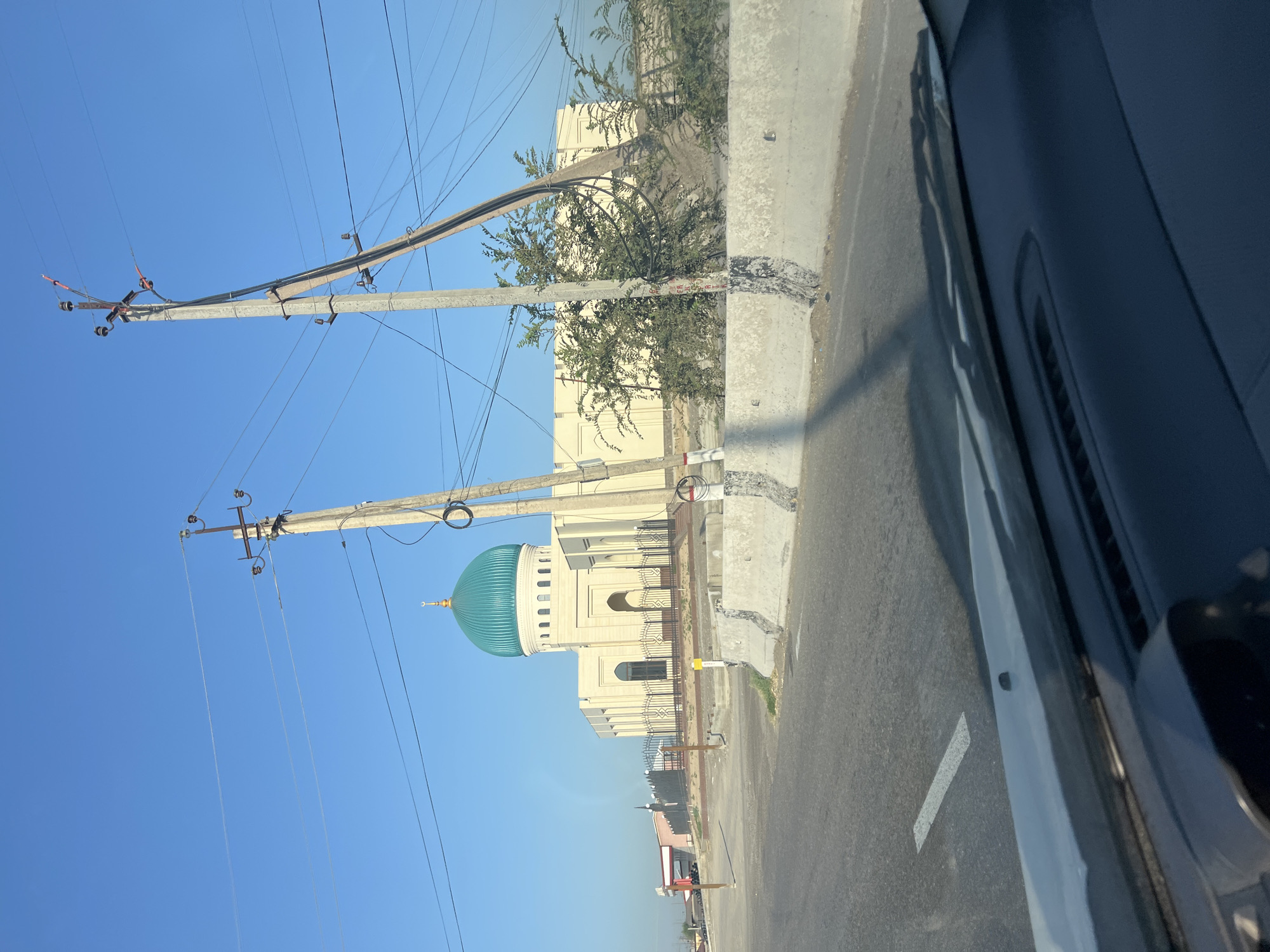 The image shows a view from inside a vehicle looking out towards a mosque with a teal-colored dome, located in Yangi Chinaz, Uzbekistan. The mosque is a light-colored, modern building with arched windows and a fence in front of it. It is surrounded by some vegetation and utility poles with many wires. The sky is clear and blue, and the road in the foreground has a white line marking. The perspective suggests the vehicle is moving on a road adjacent to the mosque.