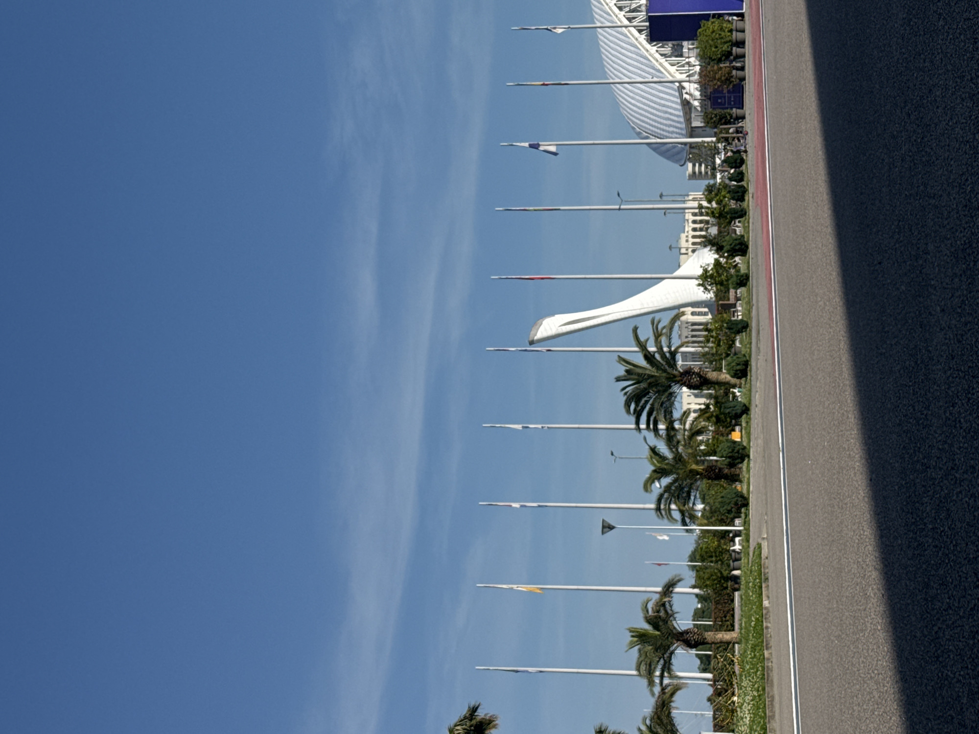The image captures an outdoor, daytime scene within the city of Sirius, Russia, specifically depicting elements of the former Sochi Olympic Park. In the mid-ground, the prominent white, stylized structure of the "Medalists' Wall," which served as the Olympic Cauldron for the Sochi 2014 Winter Olympics, is visible on the left. To its right, the distinctive, curved white roof of the Fisht Olympic Stadium dominates the skyline. A series of tall, white flagpoles, some bearing flags with various colors and designs that are not precisely identifiable, stretch horizontally across the mid-ground. Numerous green palm trees and other dense foliage are interspersed throughout the landscape, with some smaller buildings visible in the background behind the main structures. The foreground consists of an asphalt road surface, partially in shadow on the left side, featuring a clear white lane divider and a segment of red-colored pavement further back. A single person is observed riding an electric scooter on this red path. The sky is clear blue with faint, wispy cloud formations.