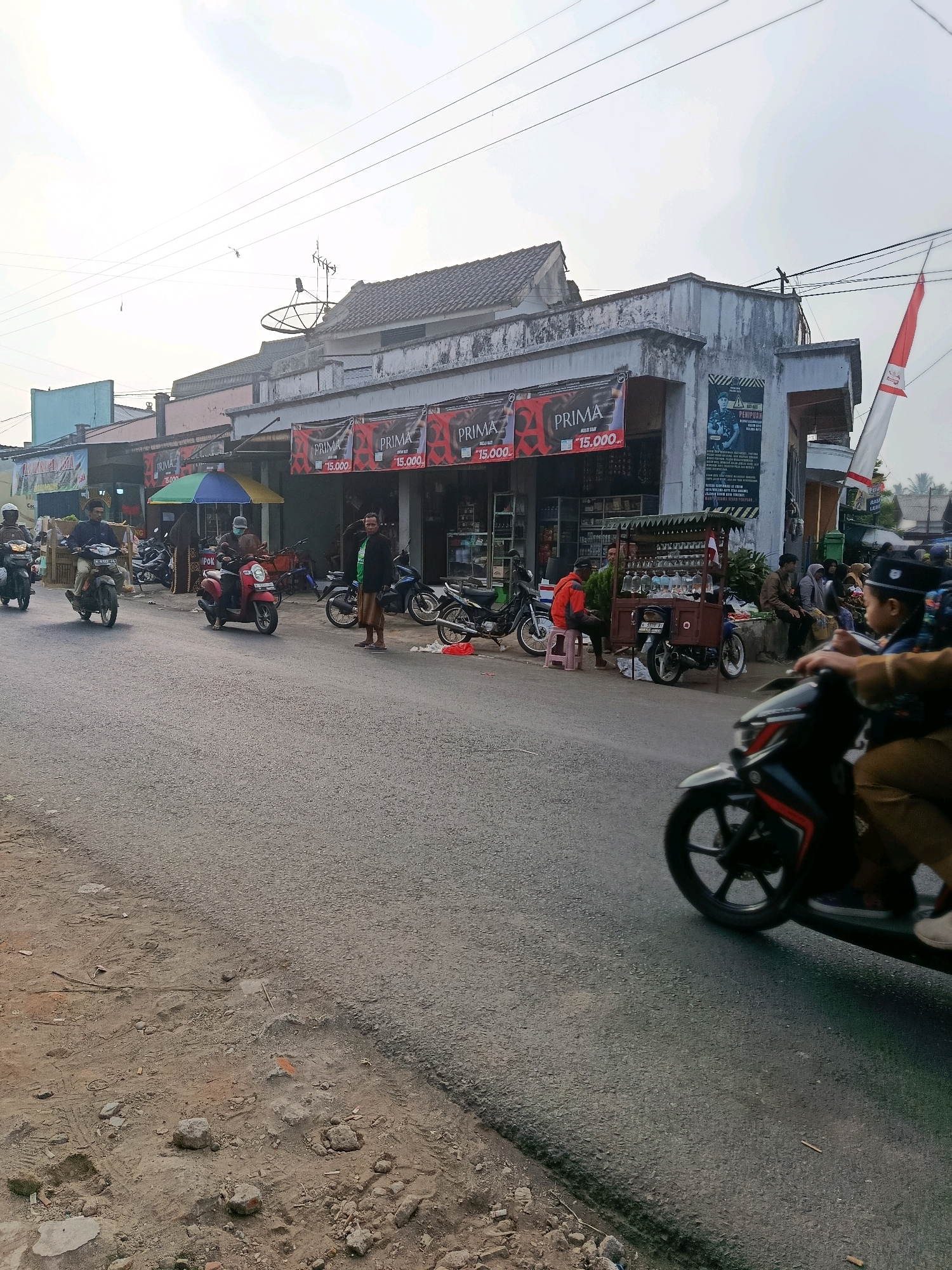 The image depicts a street scene in Tajinan, Indonesia, likely in a commercial area. A paved road runs across the foreground, with a dirt and rubble shoulder on the left. On the right side of the road, a row of low-rise buildings with awnings and signage suggests shops or small businesses. Several "PRIMA" banners with prices are visible. A red and white flag is mounted on the side of one building.
People are present in the scene, primarily associated with motorcycles. Several individuals are riding motorcycles, including one in the immediate foreground whose front wheel and lower body are blurred by motion. Other motorcycles are parked or being ridden further down the street. A street vendor is operating a cart with multiple bottles, possibly selling drinks or fuel. A small group of people are seated on a bench or curb near the vendor.
The sky is overcast with a bright, hazy quality, indicating it is daytime. Power lines are strung across the top of the image, and a satellite dish is visible on one of the building roofs. The overall impression is of a typical, active street in a local Indonesian town.
