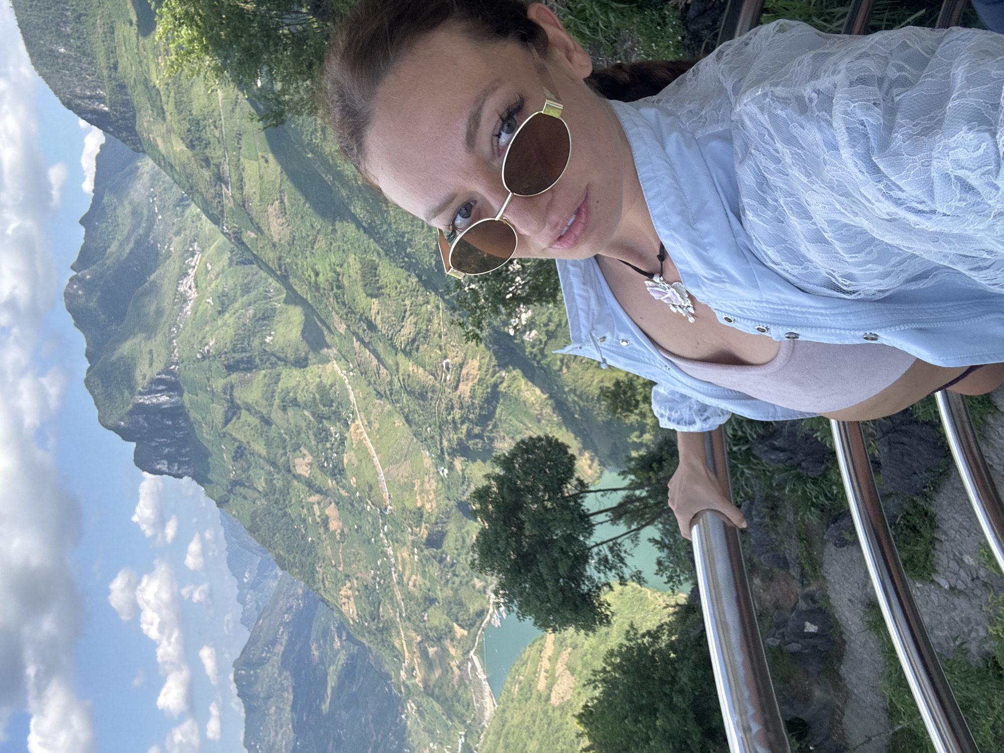 A woman in sunglasses poses for a photograph on a railing overlooking a valley in Đồng Văn Commune, Vietnam. The valley features steep, green mountains and a blue river. The woman wears a light blue shirt with lace and a necklace with a butterfly pendant. She holds onto the railing with her left hand.