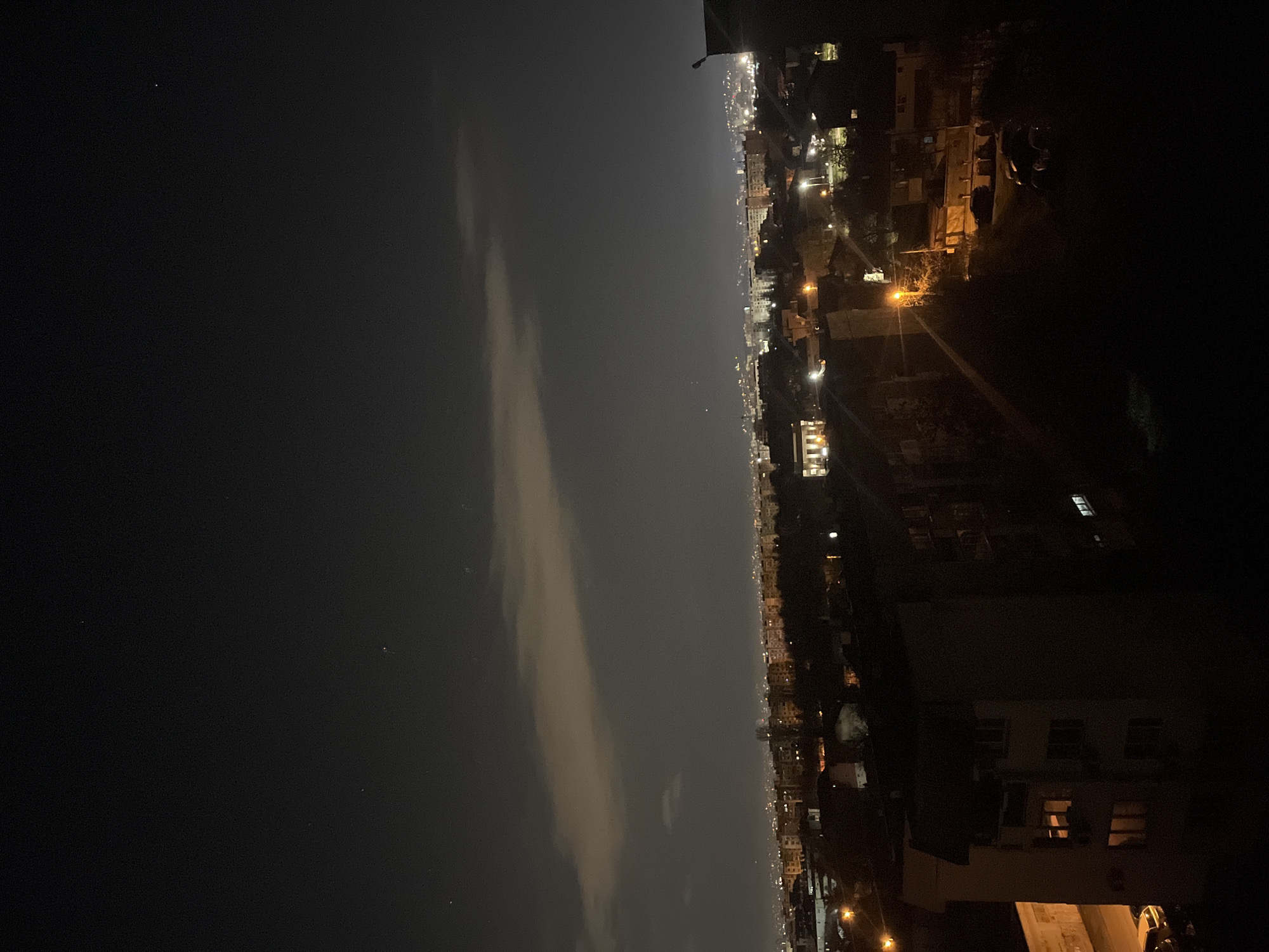 A nighttime, high-angle view shows the city of Sofia, Bulgaria, along with a dark body of water or open area to the left. Building lights and street lamps illuminate the cityscape. The left side of the image is dark, suggesting a body of water or unlit open space. A lighter cloud formation is visible in the darker space, contrasting with the city's lights. The right side of the image shows building exteriors and streetlights. The image appears to be taken from a high vantage point, offering a panoramic view of the city at night.