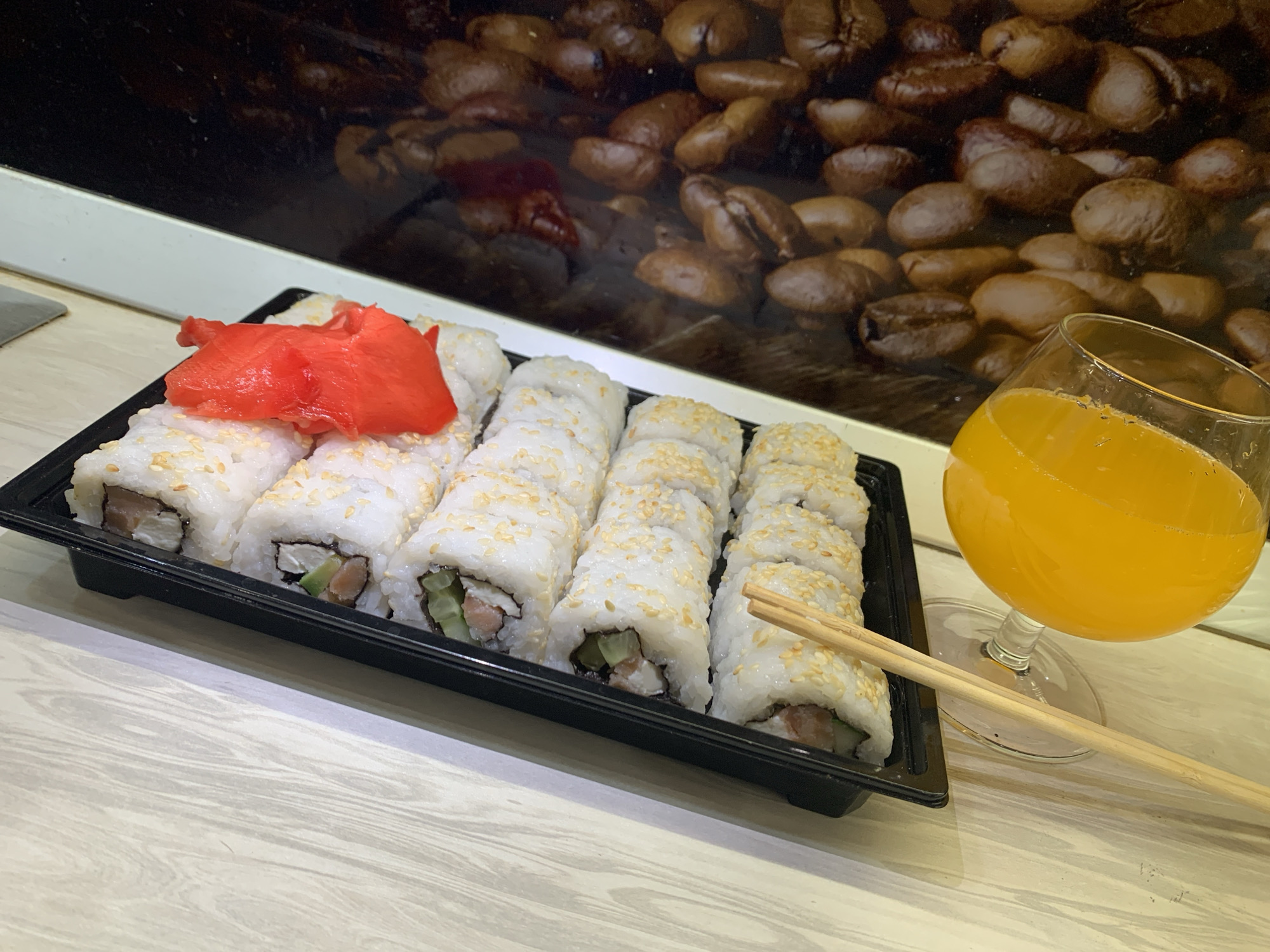 A tray of sushi rolls sits on a light-colored, possibly wooden, surface next to a glass of orange juice and a pair of chopsticks. The sushi is arranged neatly in rows and topped with sesame seeds, accompanied by a vibrant red pile of pickled ginger. The backdrop features a decorative print of coffee beans, partially obscured by a white-edged structure. The image was taken in Severodvinsk, Russia.
