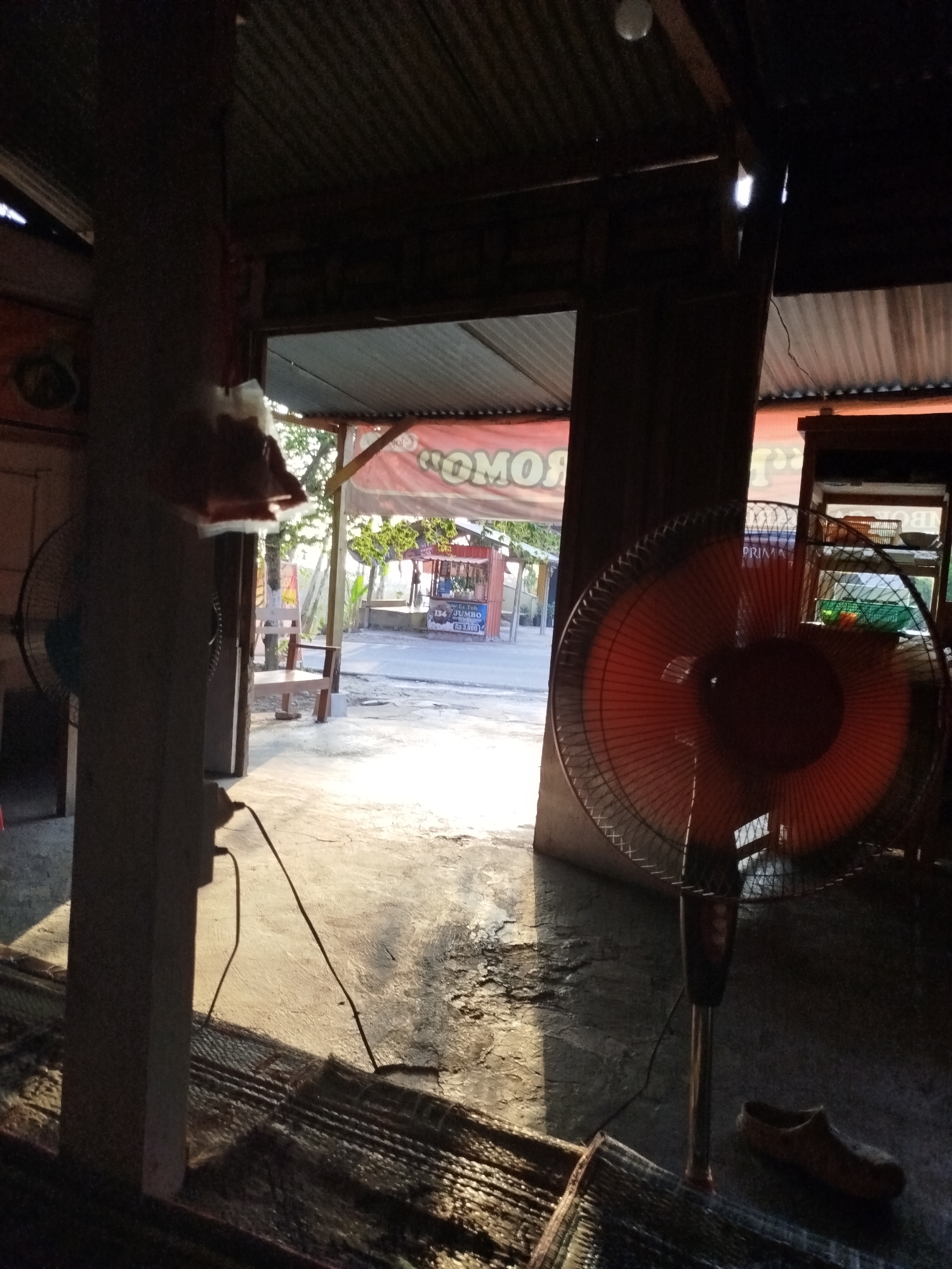 The image depicts an outdoor scene from within a covered structure, likely a roadside stall or shop in Pojok, Indonesia. The foreground is dominated by a large, orange oscillating fan in motion, casting a shadow on the concrete floor. To the left, another fan, partially obscured, is visible. A woven mat lies on the floor in front of the fans. A wooden stool or bench is positioned in the mid-ground, facing an open roadway. Beyond the bench, a red and white awning stretches across a structure with signage that reads "TOMO" and "PRIMA." Several small food or drink stalls are visible on the other side of the road, with a blue sign indicating "134 JUMBO" and a price. The environment suggests a warm, sunny day with ambient light filtering through the roof structure. A single brown sandal rests on the floor to the right of the fan.