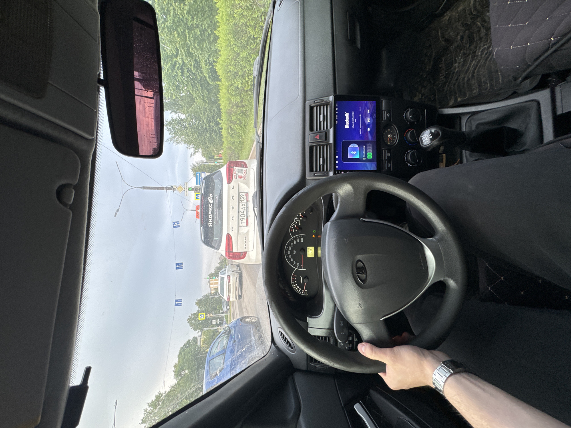 The image depicts the interior of a vehicle, specifically a Lada Priora, from the driver's perspective, looking out onto a street in the city of Perm, Russia. The driver's right hand, adorned with a metallic link-bracelet watch, rests on the steering wheel. The car's instrument cluster is visible, showing gauges for speed, RPM, fuel, and engine temperature, alongside a digital display reading "15 19" (likely time) and "6.5." Several warning lights are illuminated. An aftermarket infotainment system is installed in the center console, displaying a "Bluetooth" interface with track information "Collage (prod. Crazy-G)" by "Phono Gallery Music." A five-speed manual transmission gear shift is present.
Outside, the windshield is speckled with water droplets, indicating recent rain or mist under an overcast sky. The vehicle is stopped in traffic behind a white Lada Granta FL, which bears the Russian license plate T904AX 159, confirming its registration in Perm Krai. The rear of the Granta features a "Yandex Go" sticker, indicating a ride-sharing service, and an "ЭПОХА АВТО" (EPOCHA AUTO) sticker.
To the left of the Granta, a blue hatchback is visible. Further down the street, a white sedan is also stopped. Overhead, multiple blue square signs with two white arrows pointing upwards are suspended across the road, likely indicating lane usage or direction. A yellow triangular sign with a pedestrian icon warns of a pedestrian crossing ahead. A green traffic light is visible further down the street, and the road is lined with trees and distant urban buildings under a cloudy sky. The rearview mirror shows a reddish reflection.