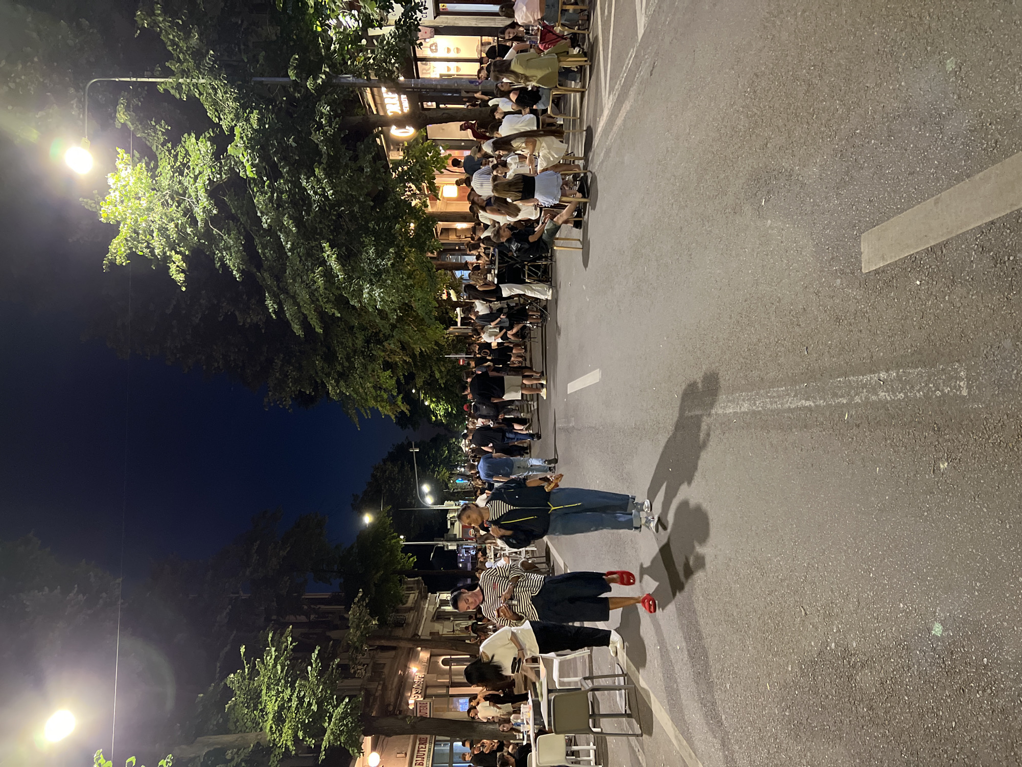 The image shows a street scene at night in Constanța, Romania, with trees lining both sides of the street and buildings visible in the background. The street is closed to traffic and has been transformed into a pedestrian zone with outdoor seating for restaurants and cafes. Numerous people are seated at tables, socializing and eating, while others walk along the street. Two individuals are walking towards the viewer in the foreground, casting long shadows on the asphalt. Streetlights illuminate the scene, creating a warm and inviting atmosphere. The overall impression is that of a lively evening in a popular urban area.