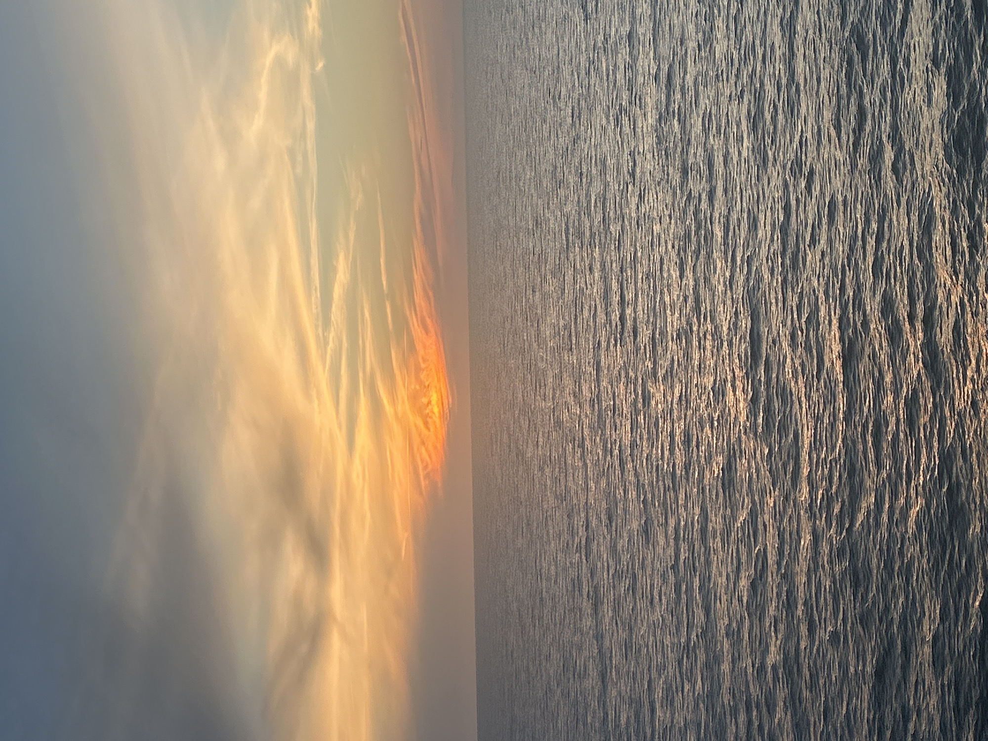 The image depicts a serene ocean view at sunset. The sky is painted with hues of orange and yellow, with wispy clouds stretching across the horizon. The sun, a bright orange orb, is low in the sky, casting a warm glow on the water. The ocean surface is calm, with gentle ripples reflecting the colors of the sky. The scene is devoid of any people or man-made objects, creating a tranquil and natural atmosphere. The location is unknown, but the scene suggests a coastal or open water setting.