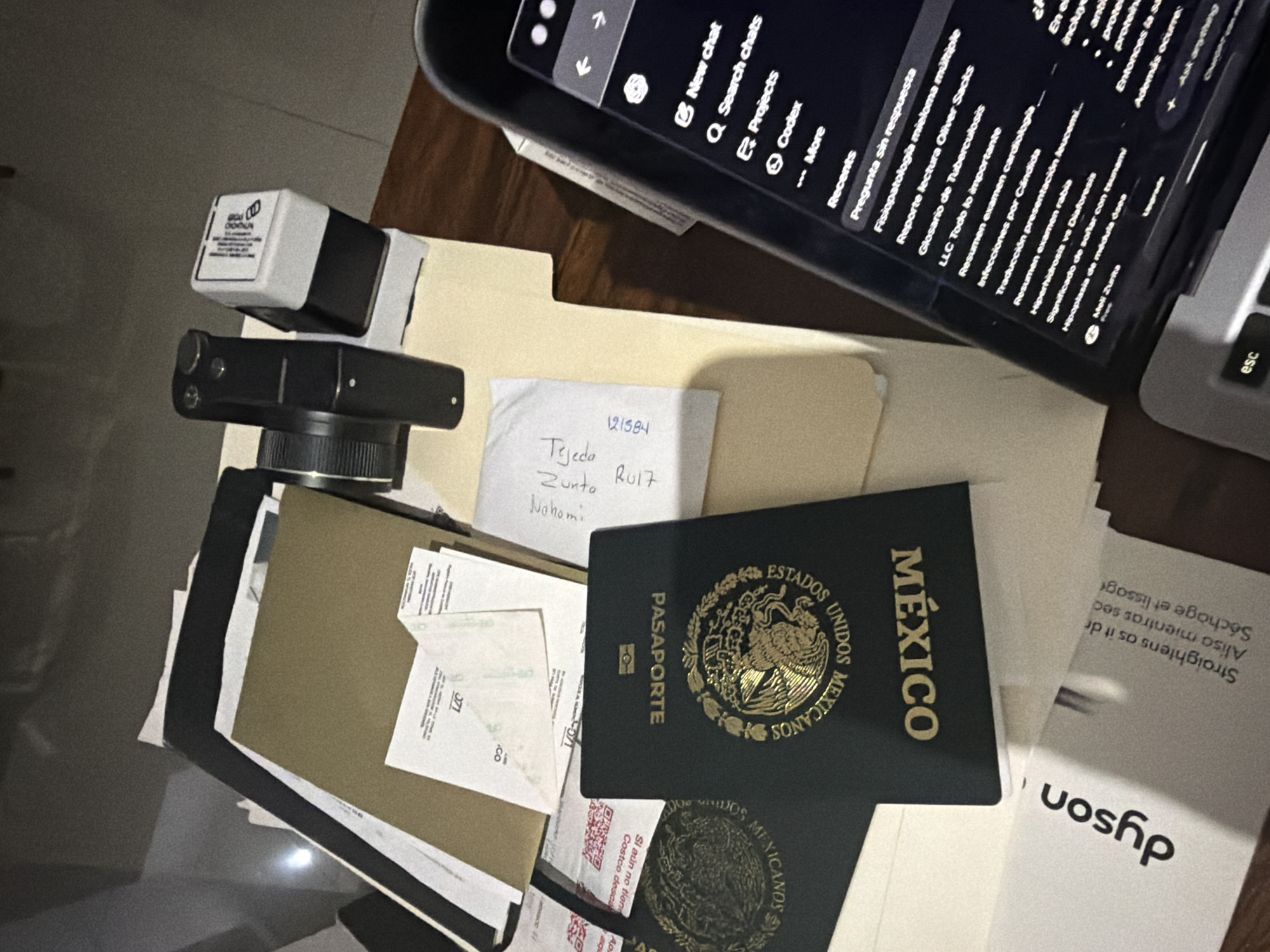 The image presents a cluttered desk scene. A laptop displays a chat interface on a dark background. Adjacent to the laptop are various objects, including a camera, a stamp, folders, documents, a passport identifying Mexico, and a Dyson brochure. A handwritten note bearing the name "Tejeda Zunto RUI7 Nohomi" is visible. The scene does not provide enough information to suggest the location is in Villahermosa, Mexico.