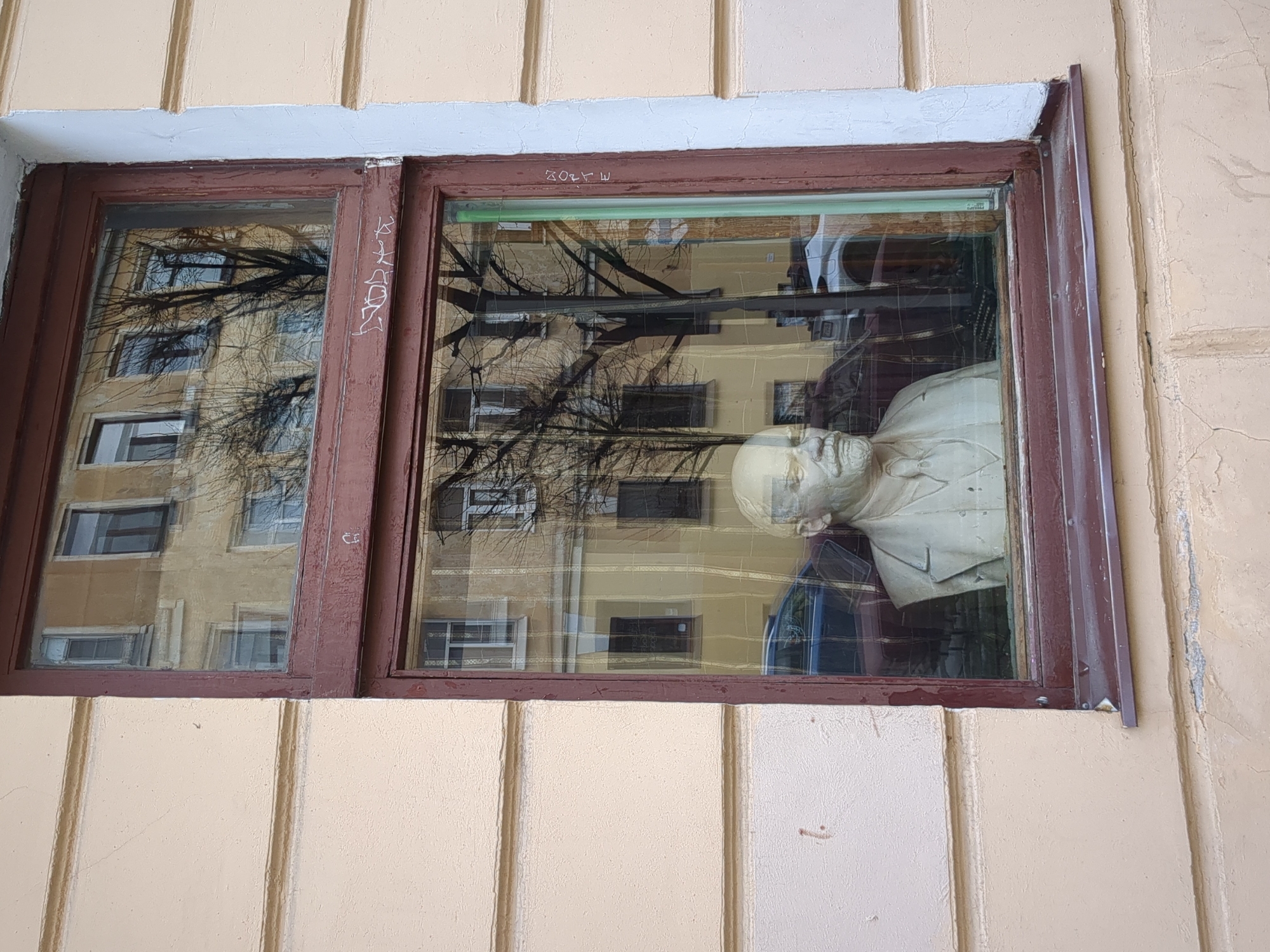 The image shows a window in a building in Saint Petersburg, Russia. The window has a dark brown frame and is divided into two panes. The reflection in the window shows a building across the street with bare trees in front. Inside the window is a bust of a man wearing a suit. The building's exterior is beige with vertical cream-colored panels. There is some graffiti on the window frame.