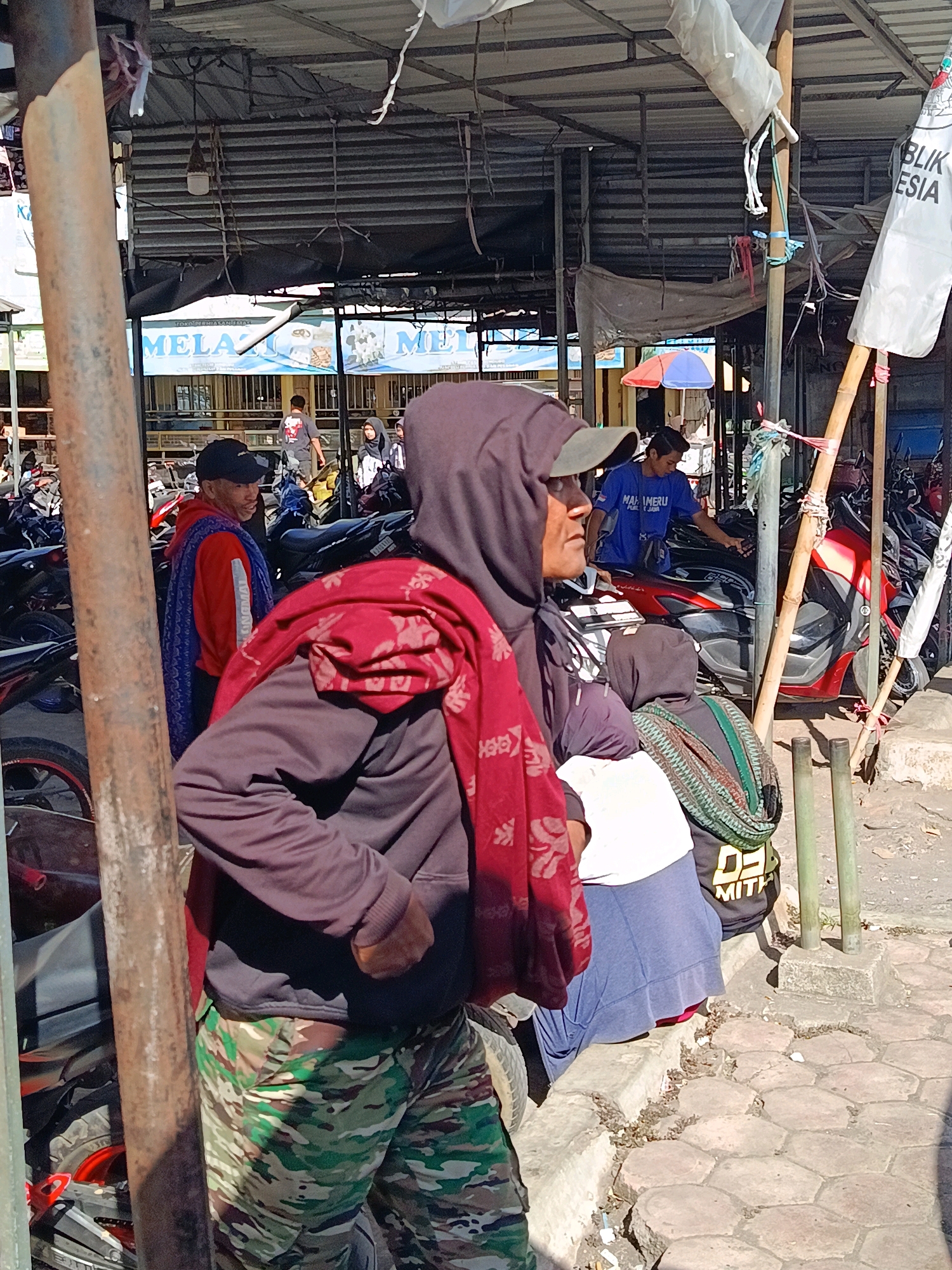 The image depicts an outdoor scene, likely a market or street, in the city of Senduro, Indonesia. In the foreground, a person wearing a maroon hooded sweatshirt, camouflage pants, and a red scarf draped over their shoulders is the primary subject. The person is standing near a metal pole and appears to be looking to their right. Behind this individual, other people are visible, including a person in a red jacket with a blue scarf, and a person wearing a blue t-shirt. Various motorcycles are parked in the background, suggesting a transportation hub or area where people frequent. A white banner with Indonesian text is visible hanging from a bamboo pole on the right side of the image. The ground is paved with hexagonal stones. The overall environment appears to be illuminated by natural sunlight, casting shadows on the ground and subjects.