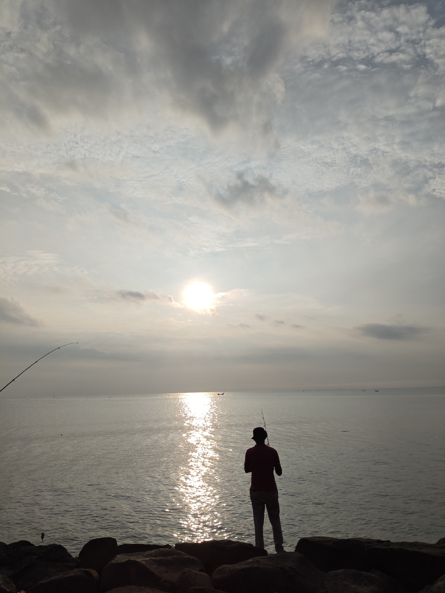 The image depicts a serene coastal scene at either sunrise or sunset in Lhokseumawe, Indonesia. The sun is low in the sky, creating a bright glare and a long, shimmering reflection across the calm waters of the sea.
The main subject is a person, seen in silhouette from behind, standing on dark rocks along the shore. The person appears to be male, wearing a hat and a shirt, and is holding a fishing rod, actively engaged in fishing. Another fishing rod is partially visible on the left side of the frame, arcing over the water.
The sky above is partly cloudy, with a mix of darker clouds and brighter patches where the light filters through. The horizon extends far into the distance, where several small boats are faintly visible on the water. The foreground is dominated by large, dark rocks, providing a stable platform for the fisherman. The overall atmosphere conveys peace, solitude, and the quiet patience of fishing at the end or beginning of the day. There is no visible text in the image.