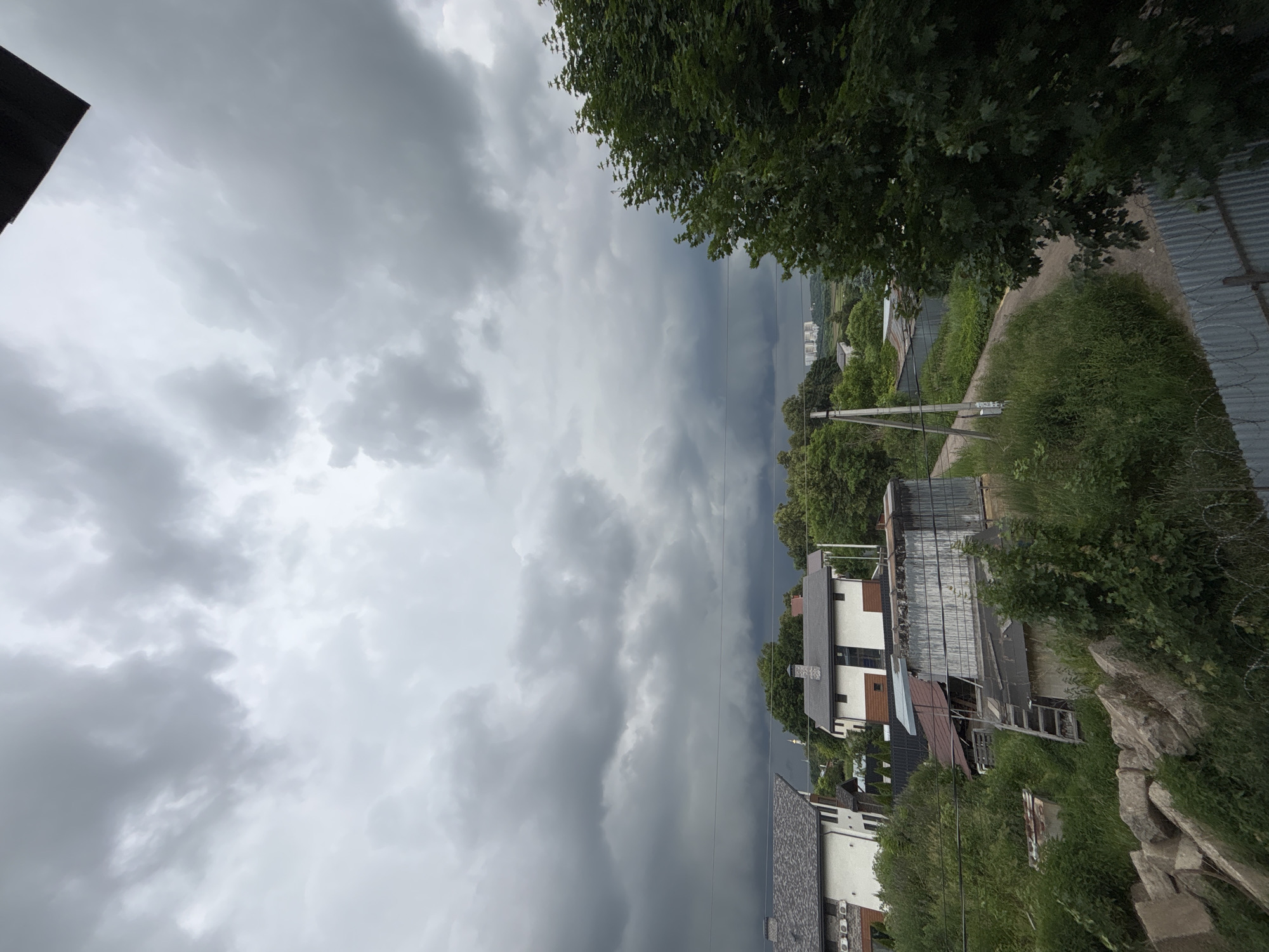 A low-angle, wide shot captures a cloudy sky above a rural residential area. In the foreground, a large evergreen tree dominates the right side of the frame, its leaves a deep green against the overcast sky. To the left, several houses with grey, peaked roofs are visible, nestled amongst lush green foliage. A dirt road winds through the scene, bordered by tall grass and what appear to be sheds or outbuildings with corrugated metal roofs. In the far distance, a cluster of modern, multi-story buildings can be seen on the horizon. The overall impression is that of an impending storm, with heavy, dark clouds gathering overhead. The scene appears to be located in Ostrov, Russia.