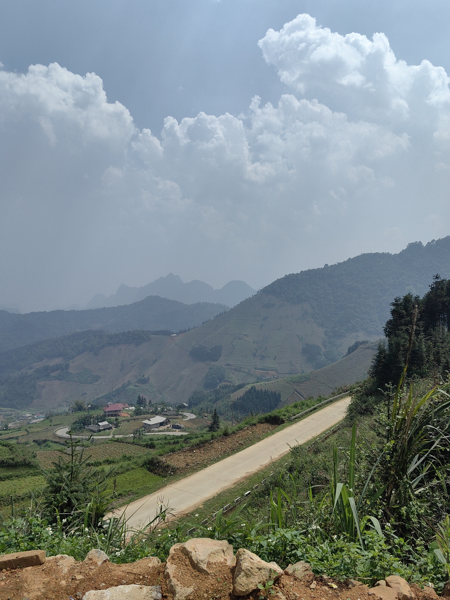 A daytime shot in Xã Cần Yên, Vietnam, shows a winding road cutting through a lush, mountainous landscape under a cloudy sky. The road is light-colored concrete or asphalt. The terrain consists of green hills and terraced farmland. In the background, hazy mountain ranges are visible, diminishing in clarity with distance. The foreground features roadside vegetation and exposed soil and rocks. A small village with several buildings is nestled amongst the hills in the middle distance.